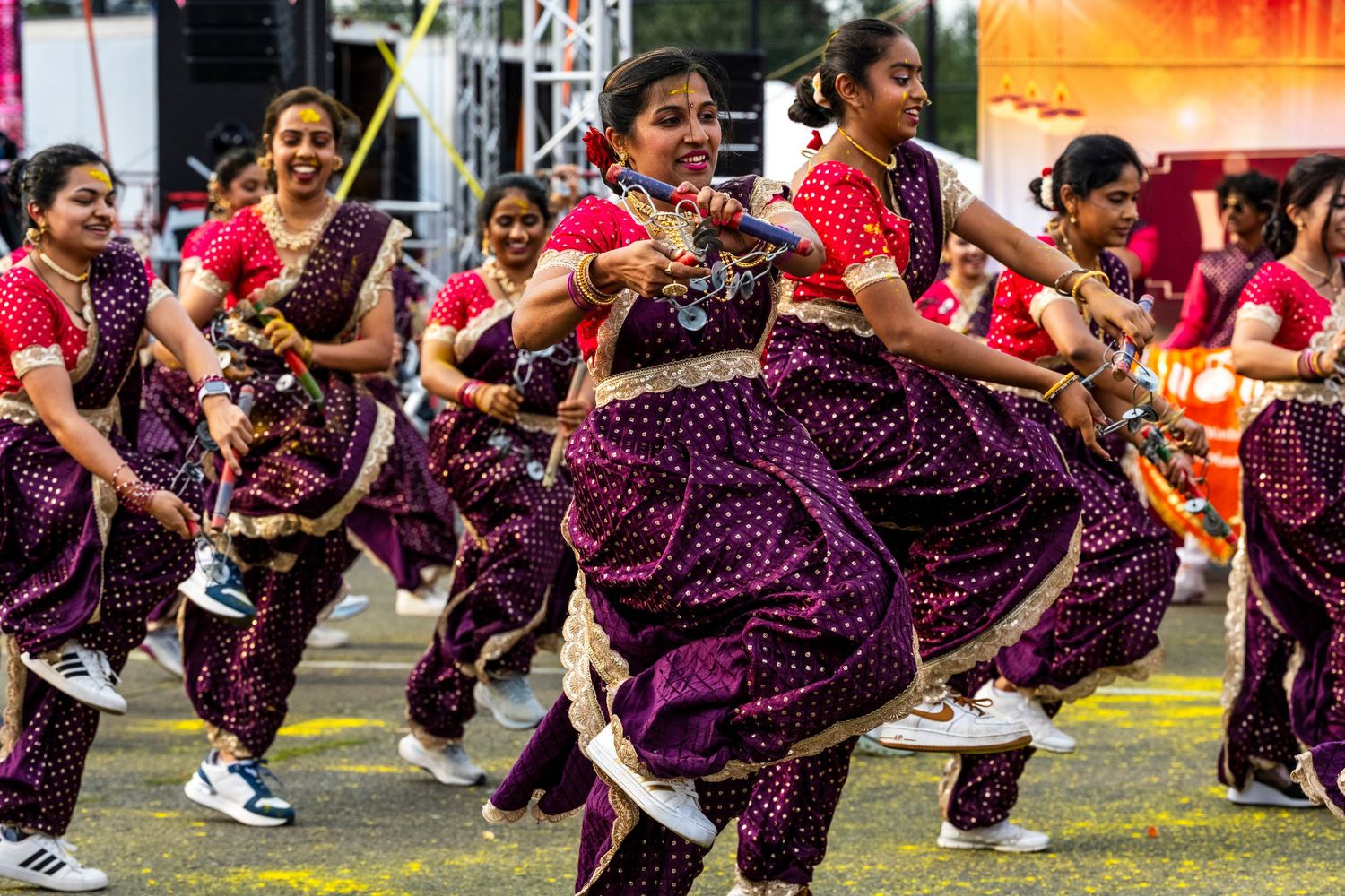 A group of women in purple sarees perform traditional Indian folk dance at an outdoor cultural festival.