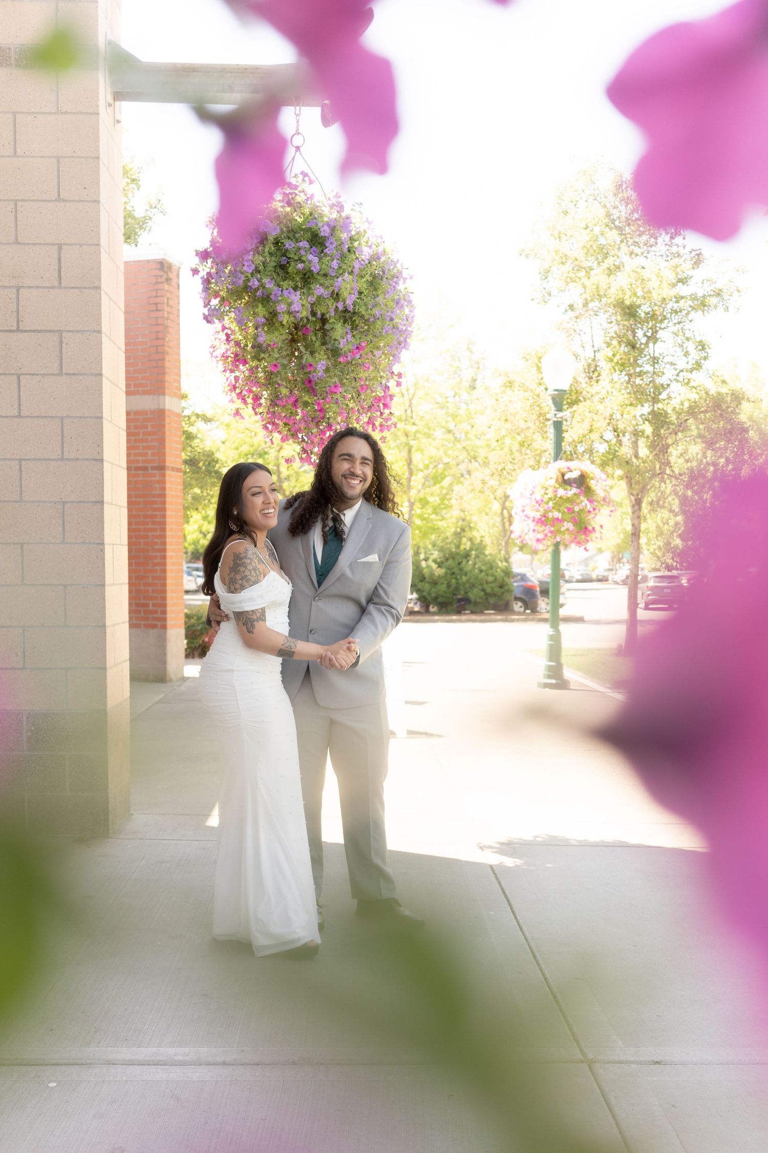 Smiling bride and groom embrace on a sunny sidewalk with colorful hanging flower baskets overhead.