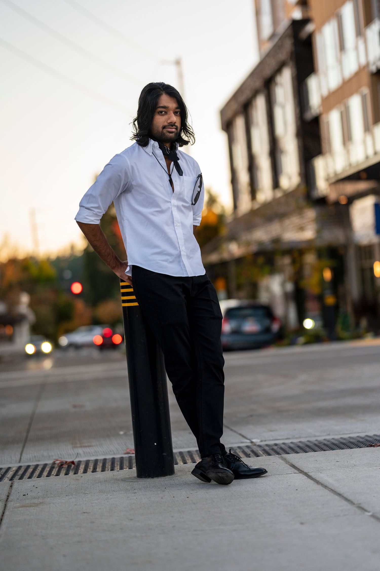 Man in white shirt and black pants posing on urban sidewalk at dusk.