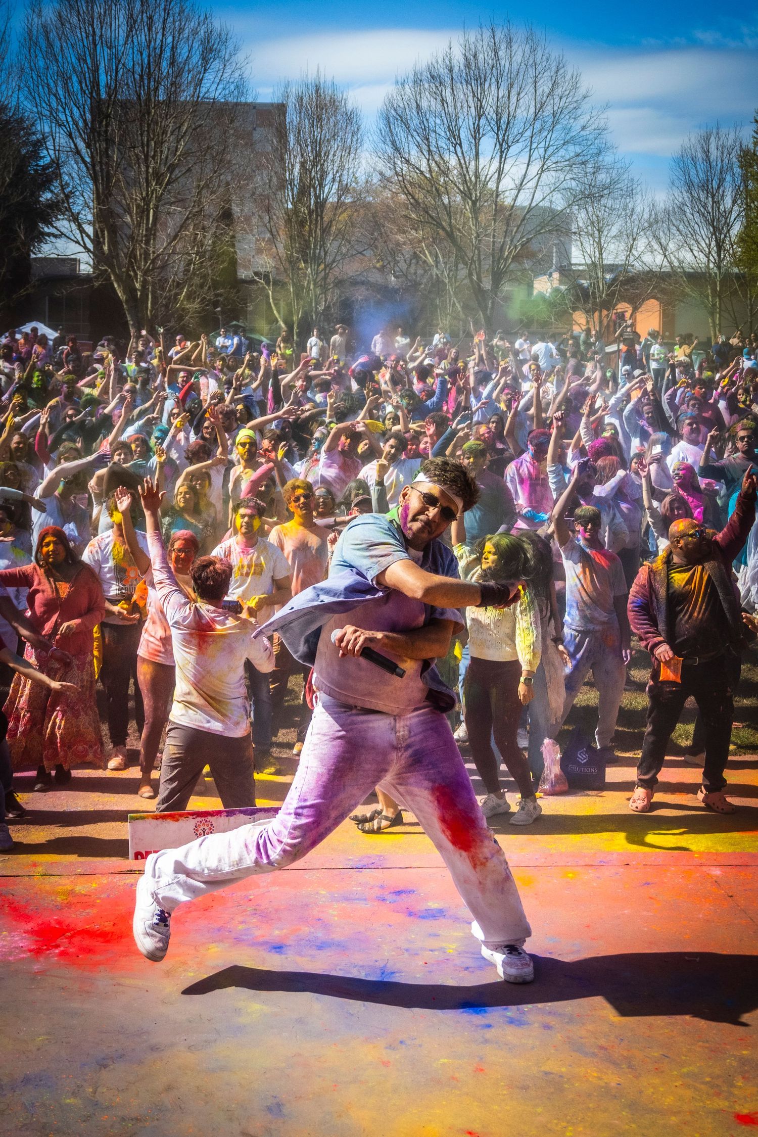 Joyful man lifting friend during colorful Holi festival celebration with crowd in background.