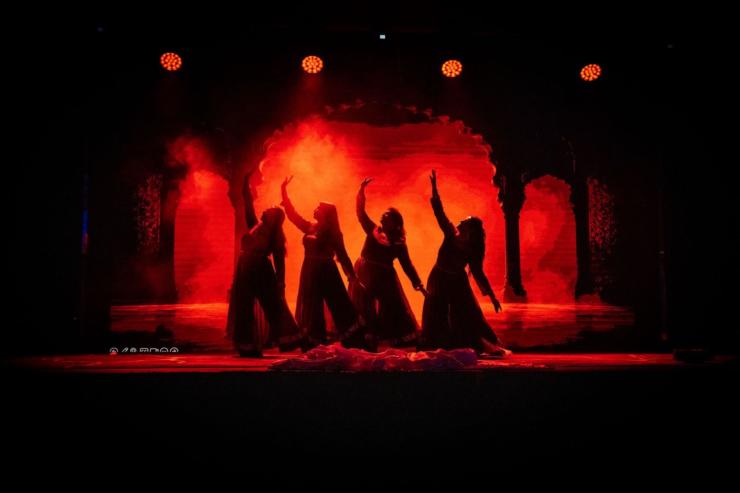 Four dancers silhouetted against vibrant red smoke lighting on a theatrical stage.