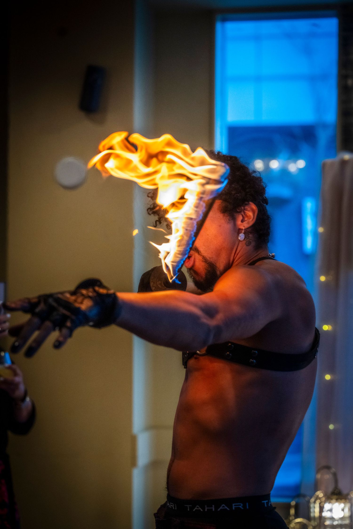 Shirtless fire breather performing dramatic flame stunt at indoor event.