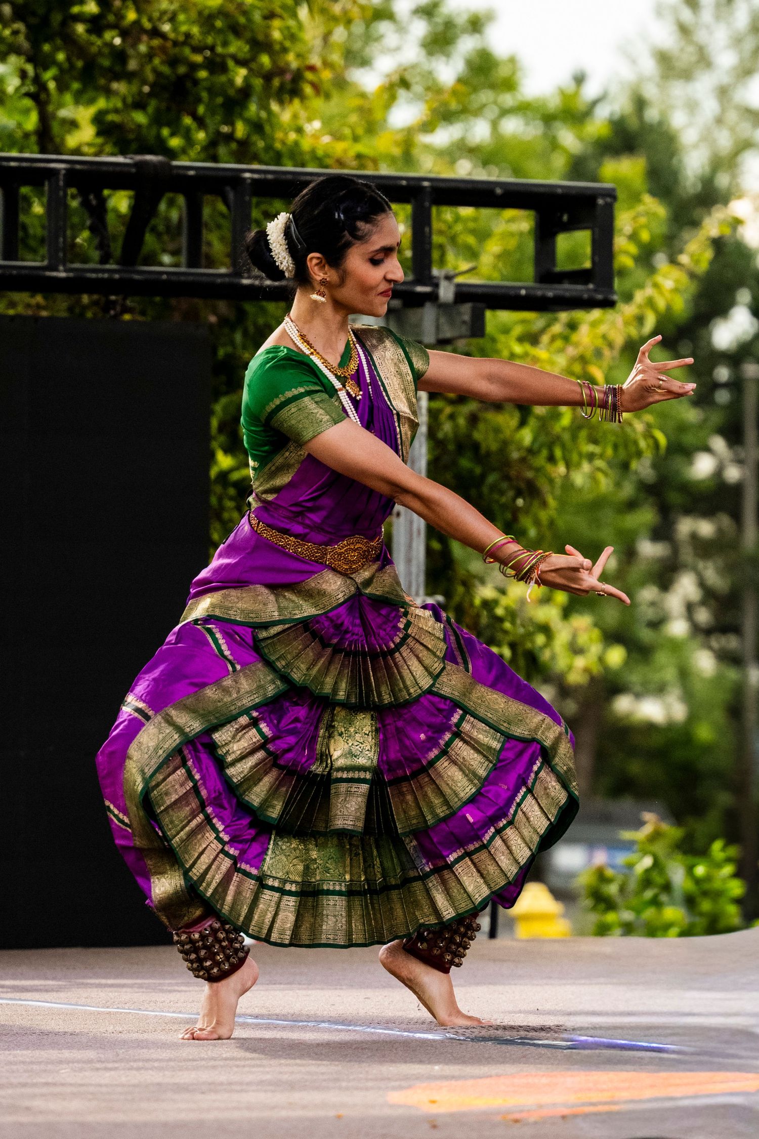 Female Bharatanatyam dancer in purple and gold saree performing outdoors on stage.