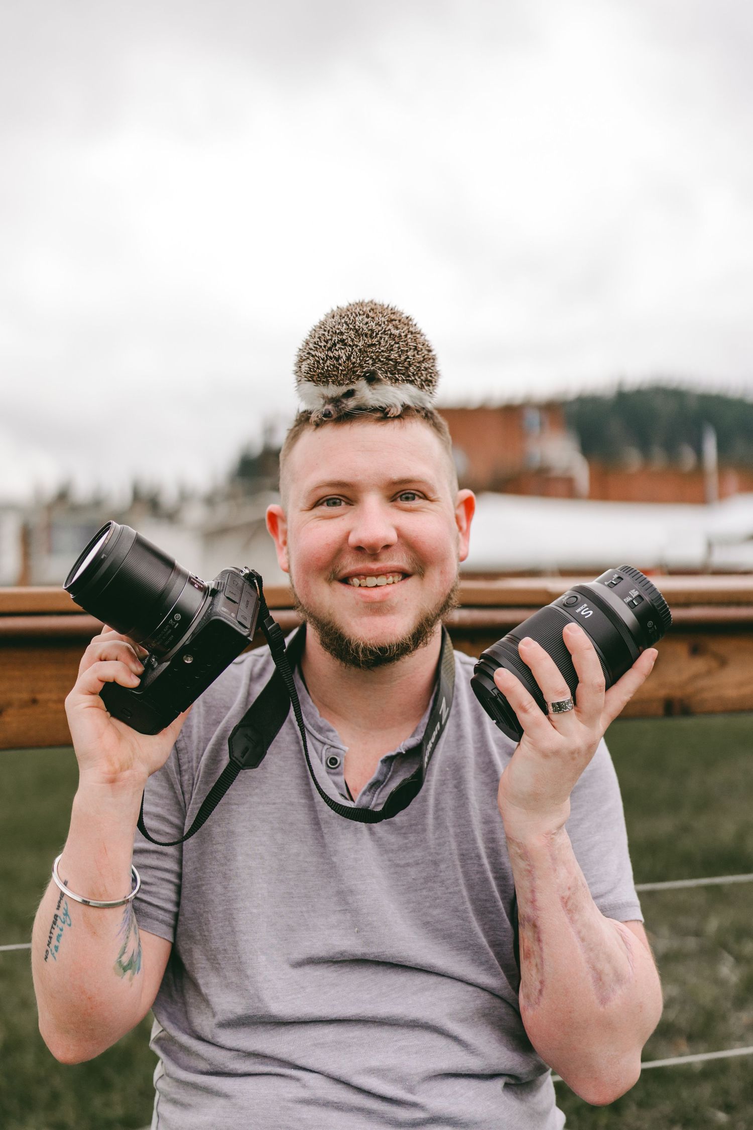 Smiling photographer holding two cameras with a hedgehog sitting on his head outdoors.
