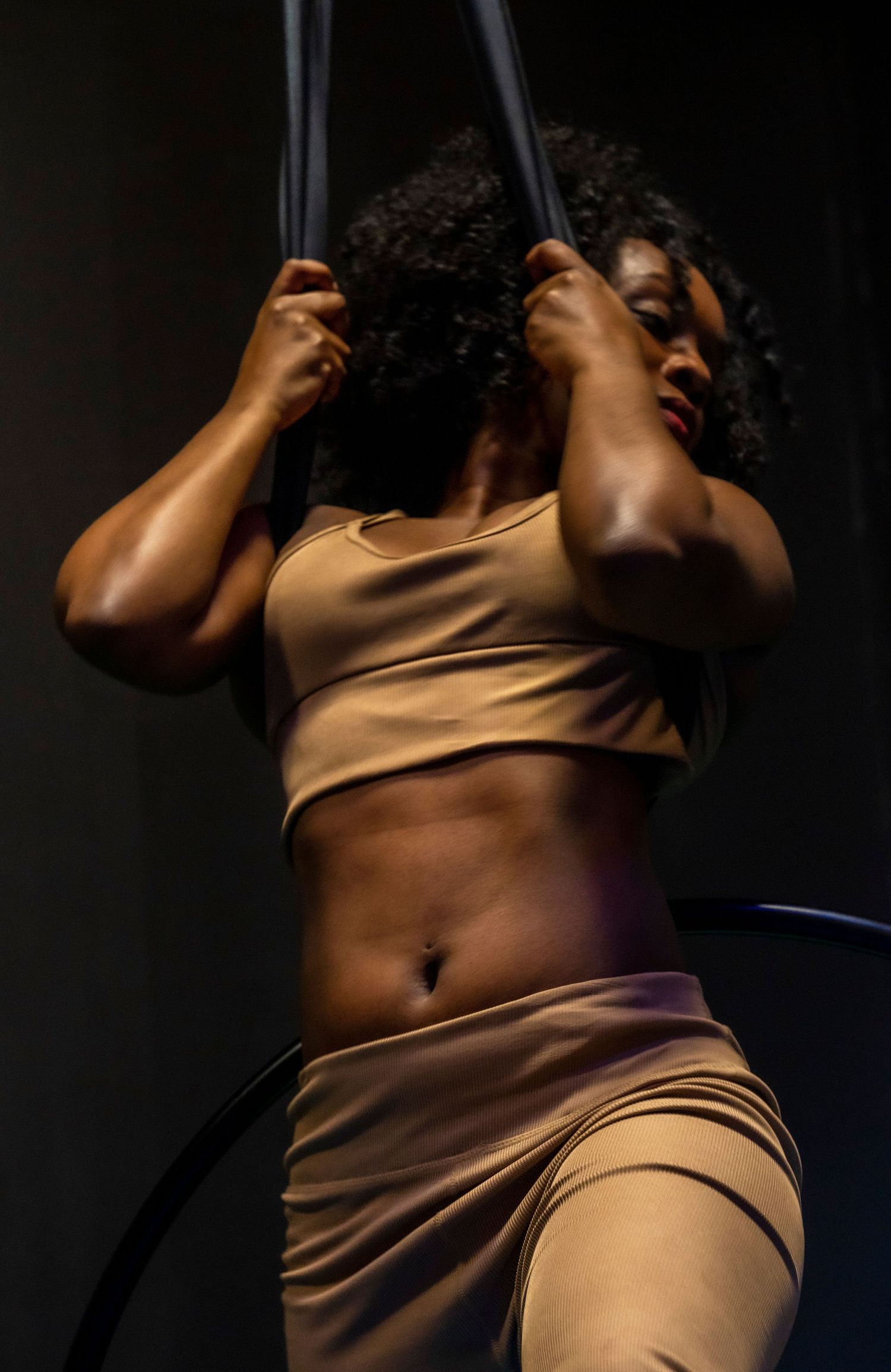 A woman in a tan crop top and skirt holds aerial silks in a dramatic, dimly lit studio setting.