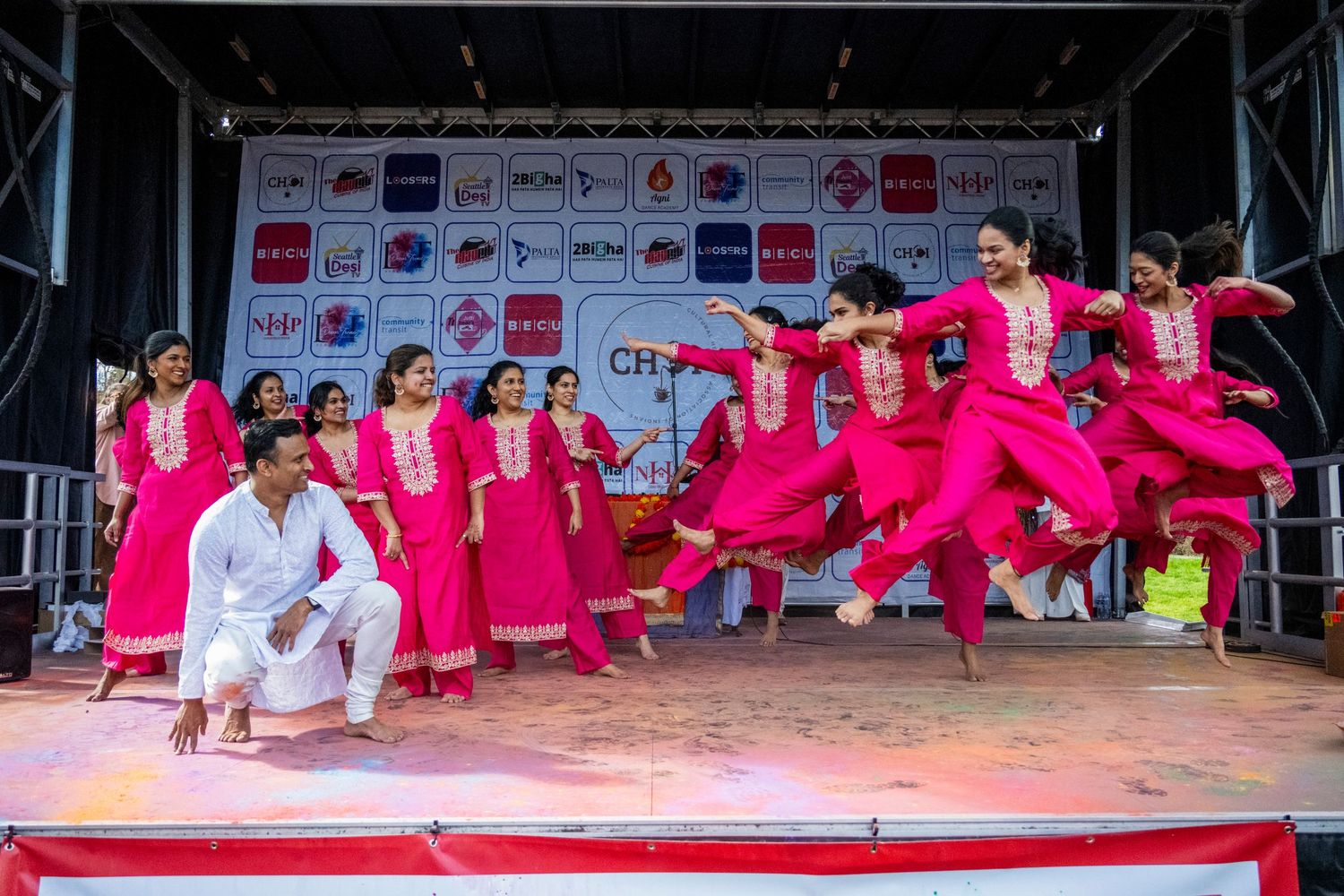 Pink-clad Bollywood dance troupe performing energetic jump sequence on outdoor festival stage.