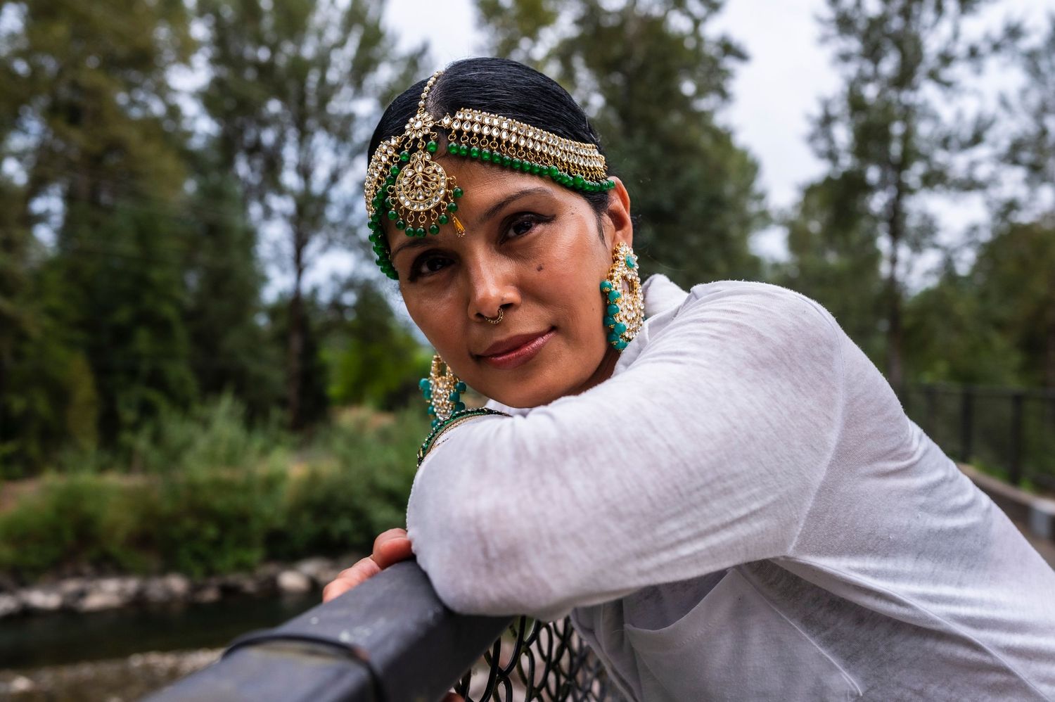 Woman in traditional Indian jewelry and headpiece poses outdoors near greenery.