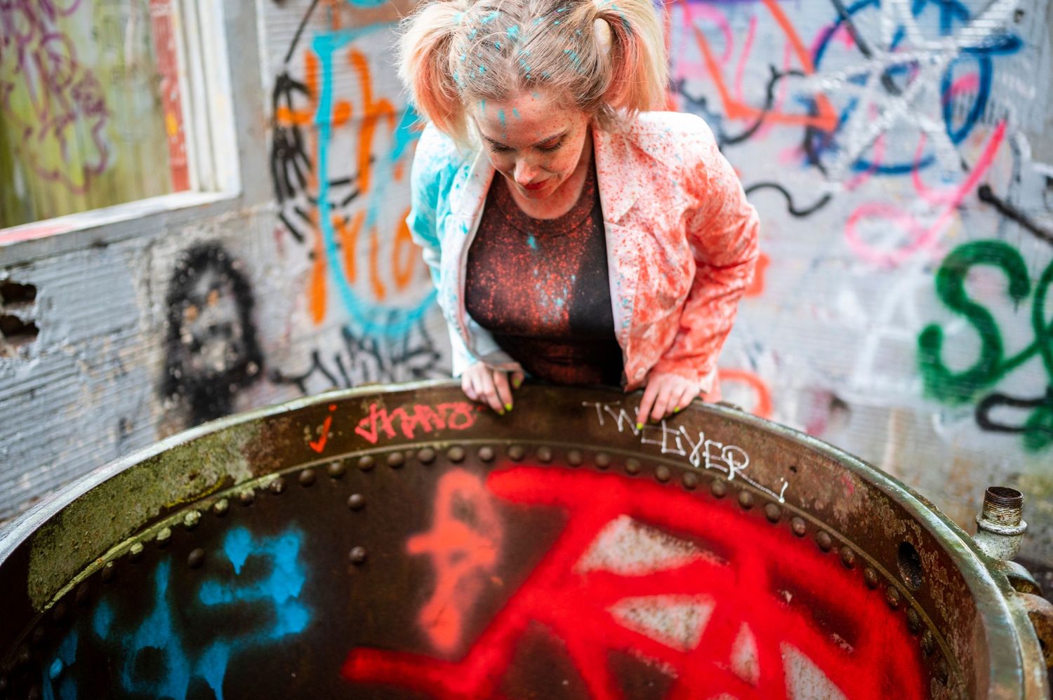 Woman with pigtails leaning over a graffiti-covered metal barrel in a colorful street art setting.