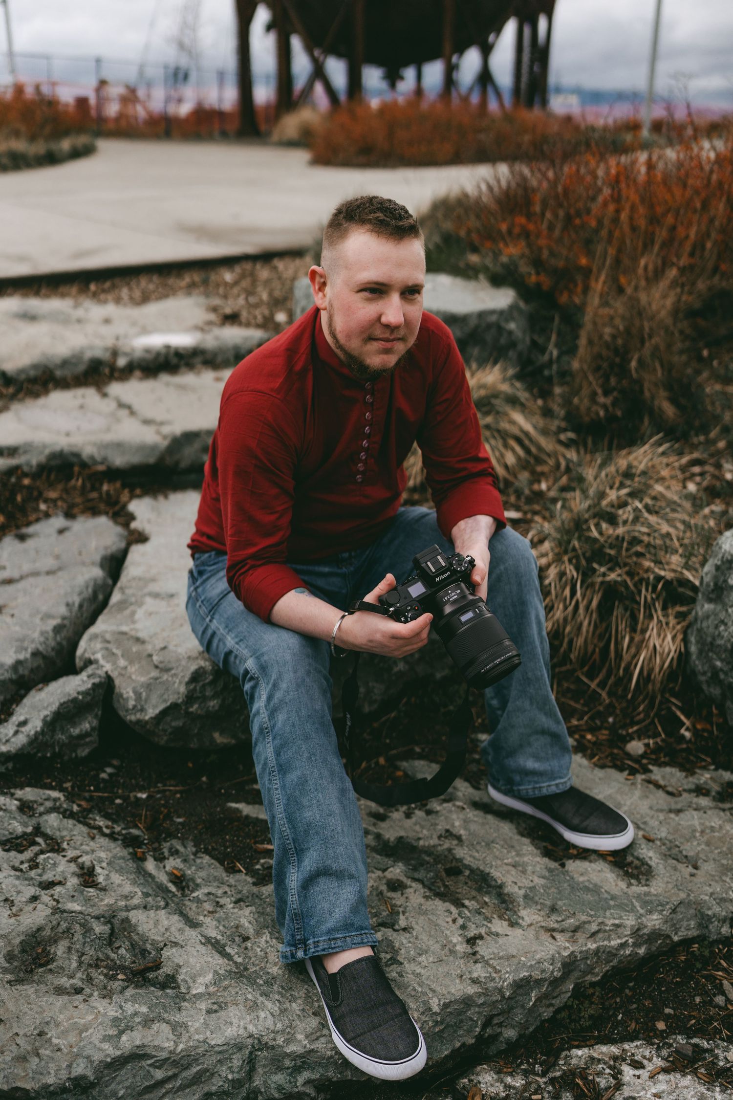 Man in red sweater sitting on rocks holding a camera at an outdoor location.