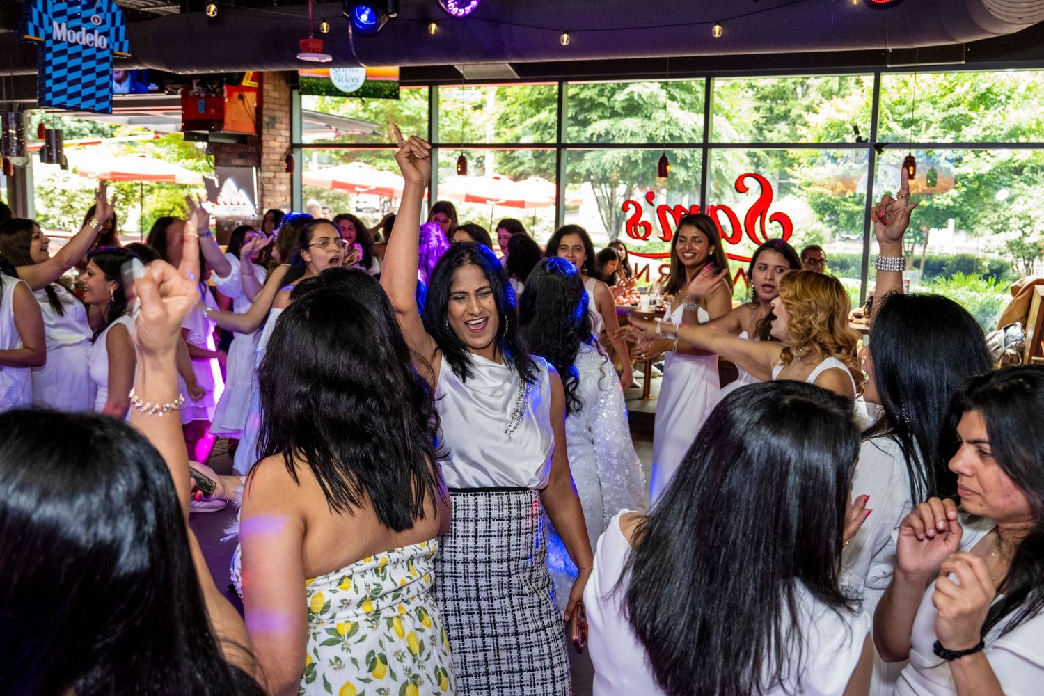 A lively crowd of women in white dance and celebrate together inside a festive bar venue.