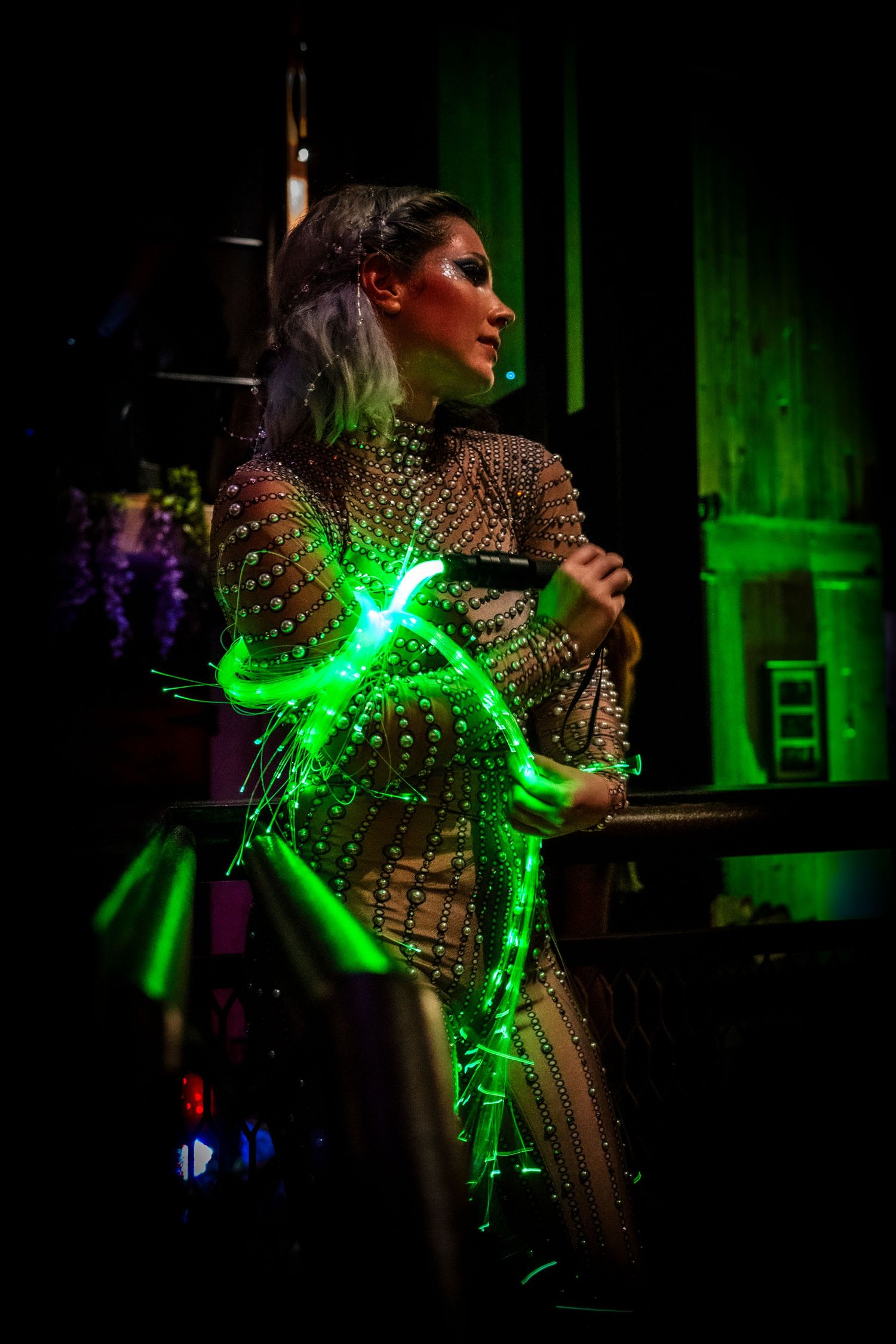 A woman in a rhinestone bodysuit holds glowing green light props on a dimly lit indoor stage.