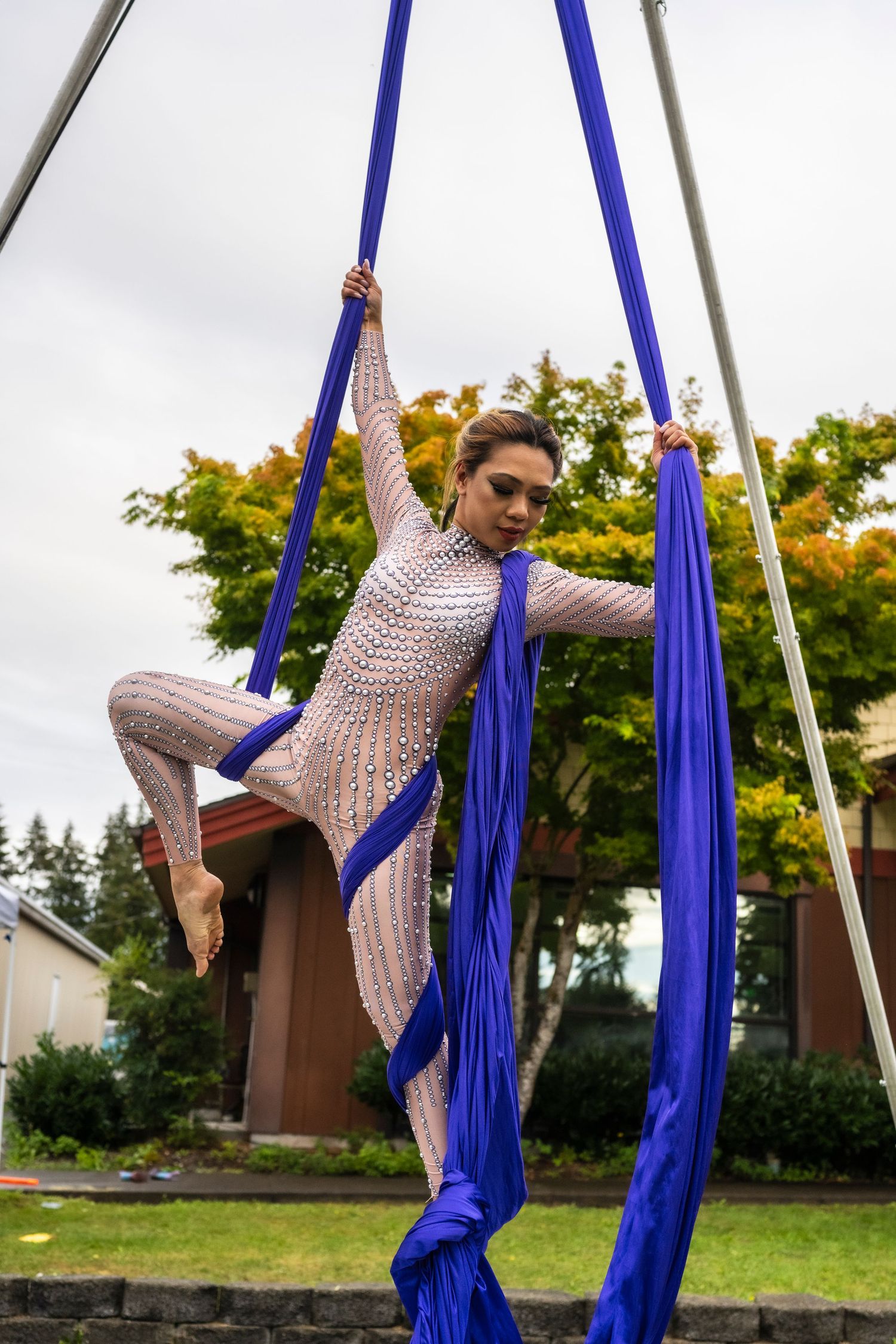 A female aerialist performs gracefully on blue aerial silks outdoors against a cloudy sky background.