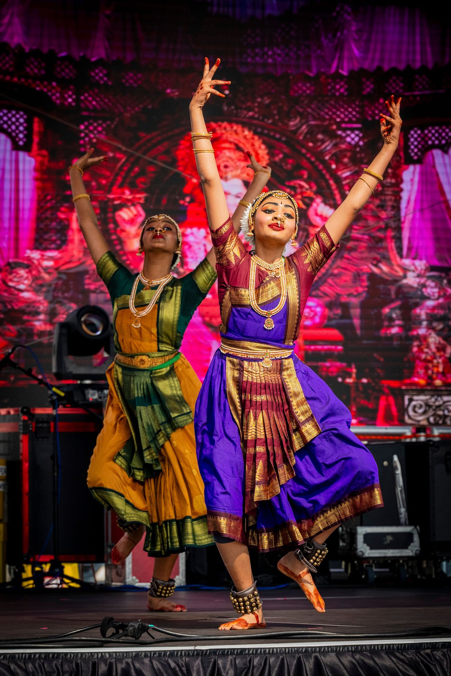 Two young girls in colorful Bharatanatyam costumes performing classical Indian dance on stage.