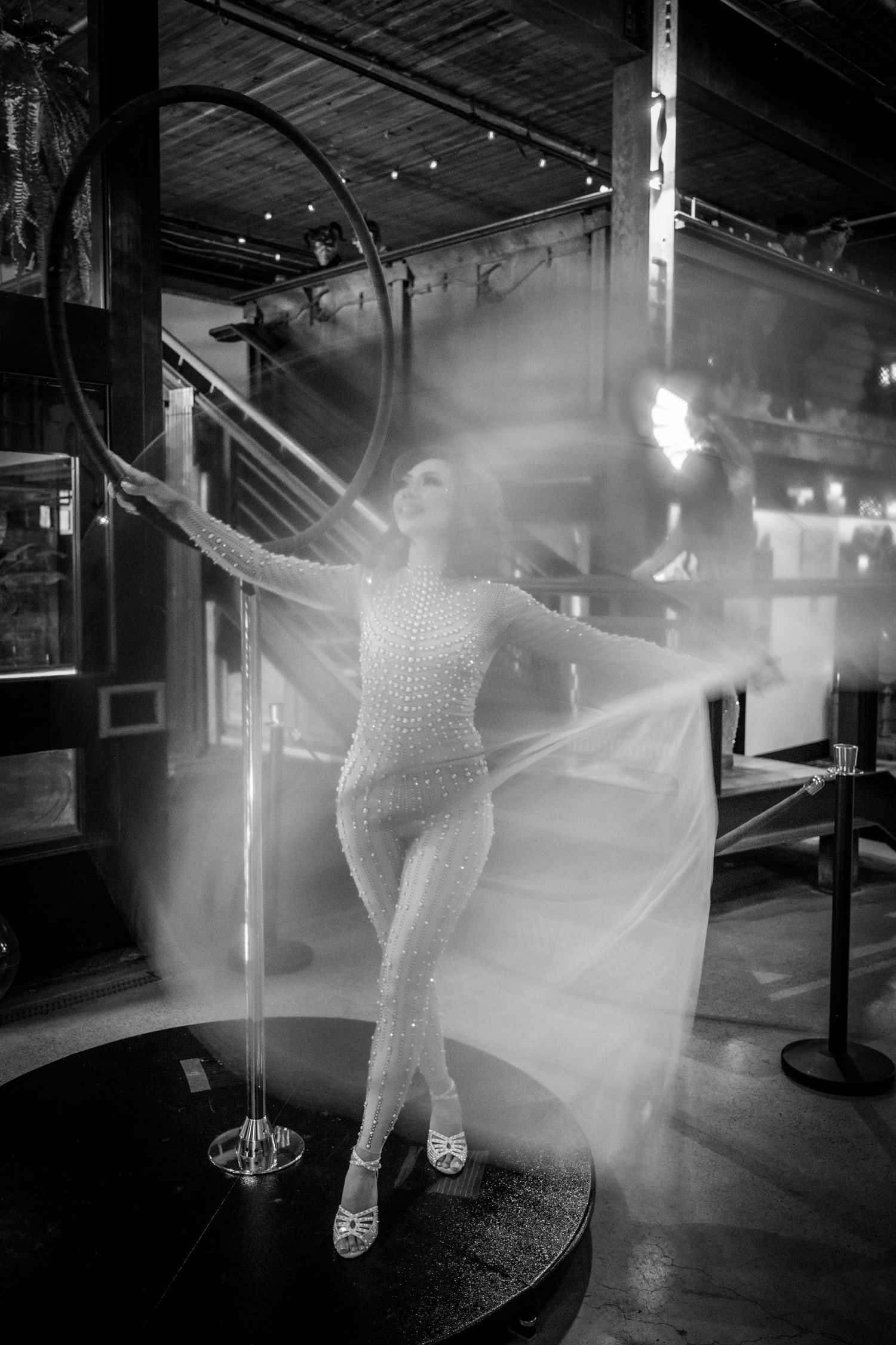 A woman in a sequined bodysuit poses dramatically with flowing fabric and fire in a black and white photo.