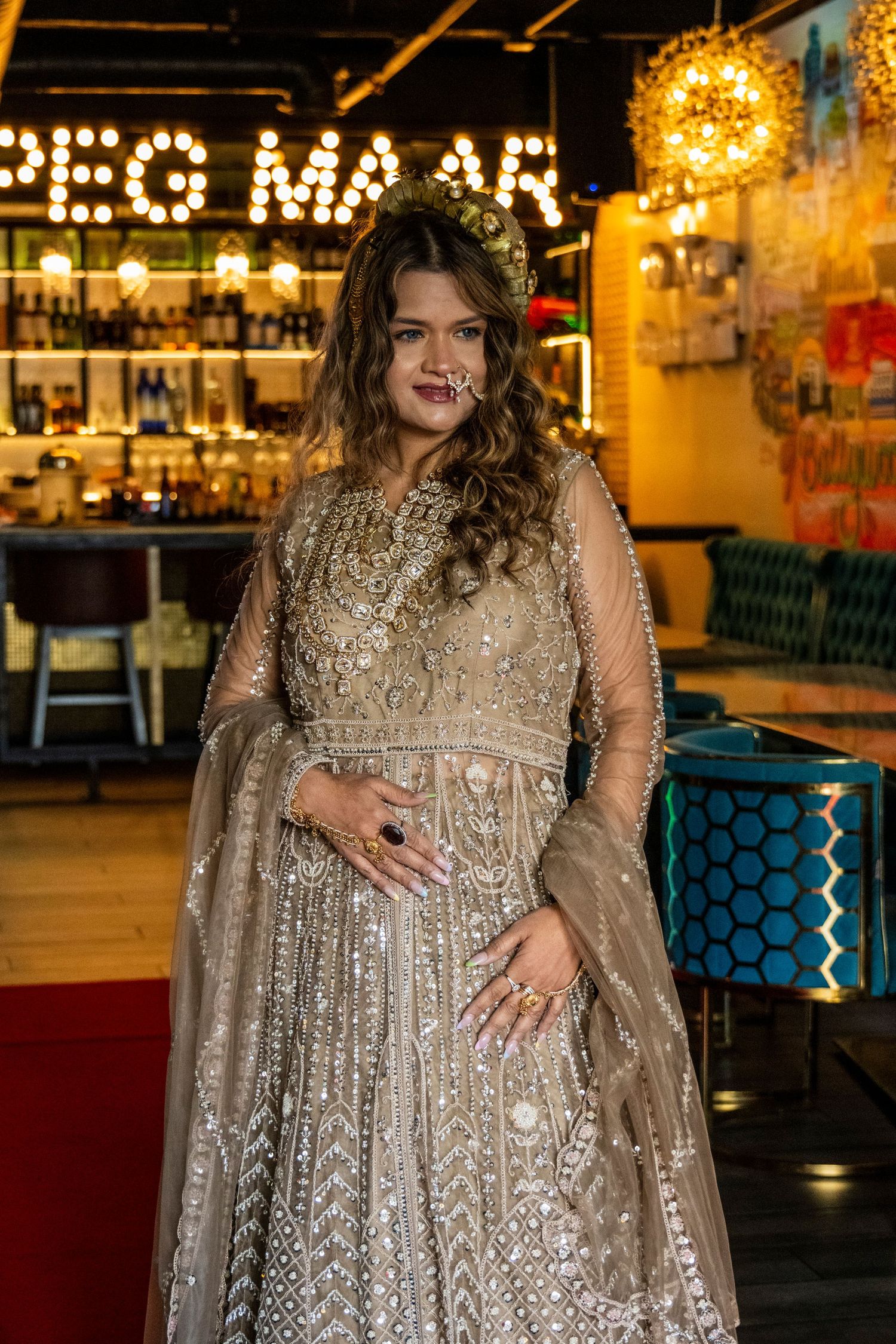 Woman in ornate gold Indian bridal lehenga with dupatta posing in front of lit bar.