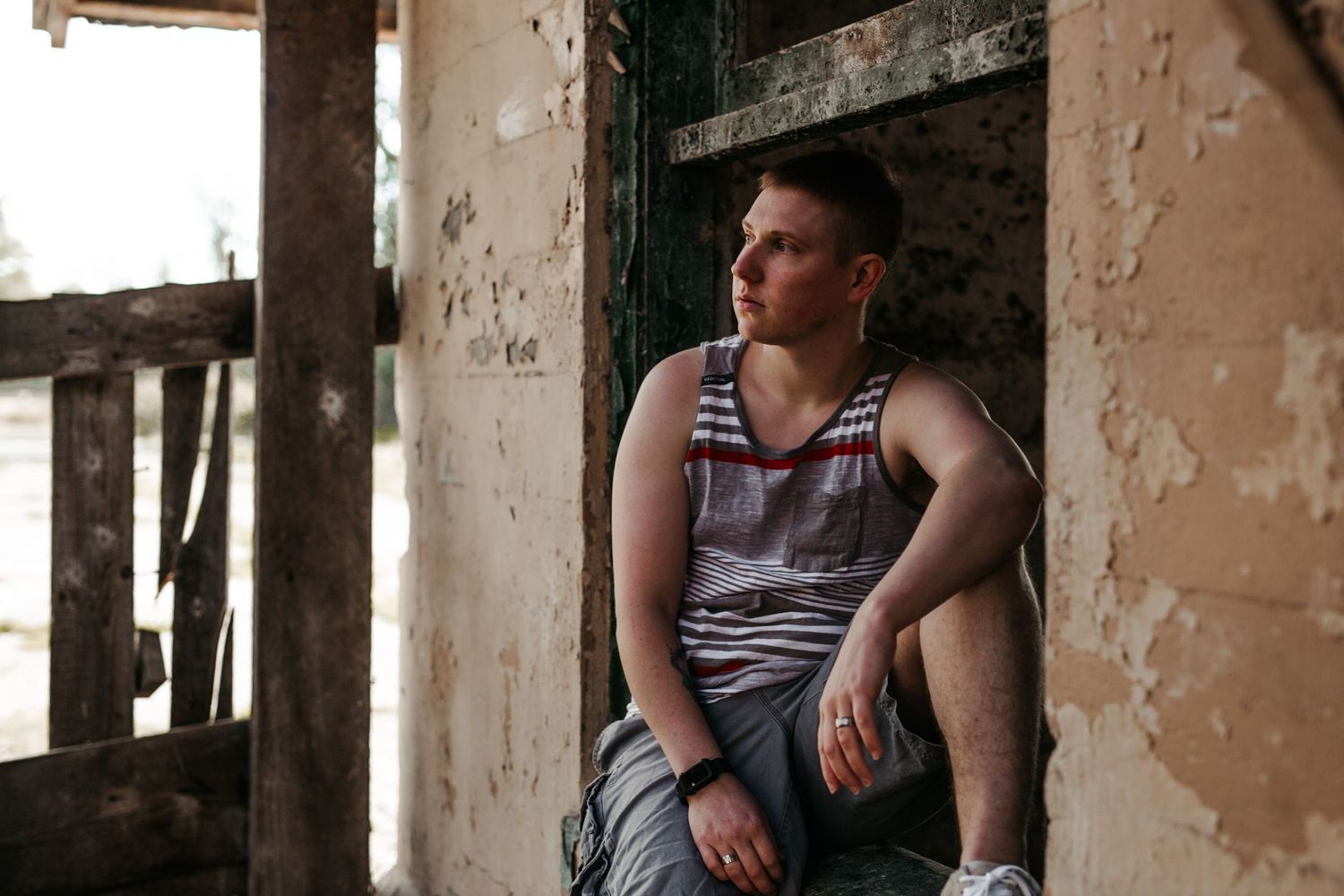 Young man in striped tank top sitting in a rustic abandoned building doorway.