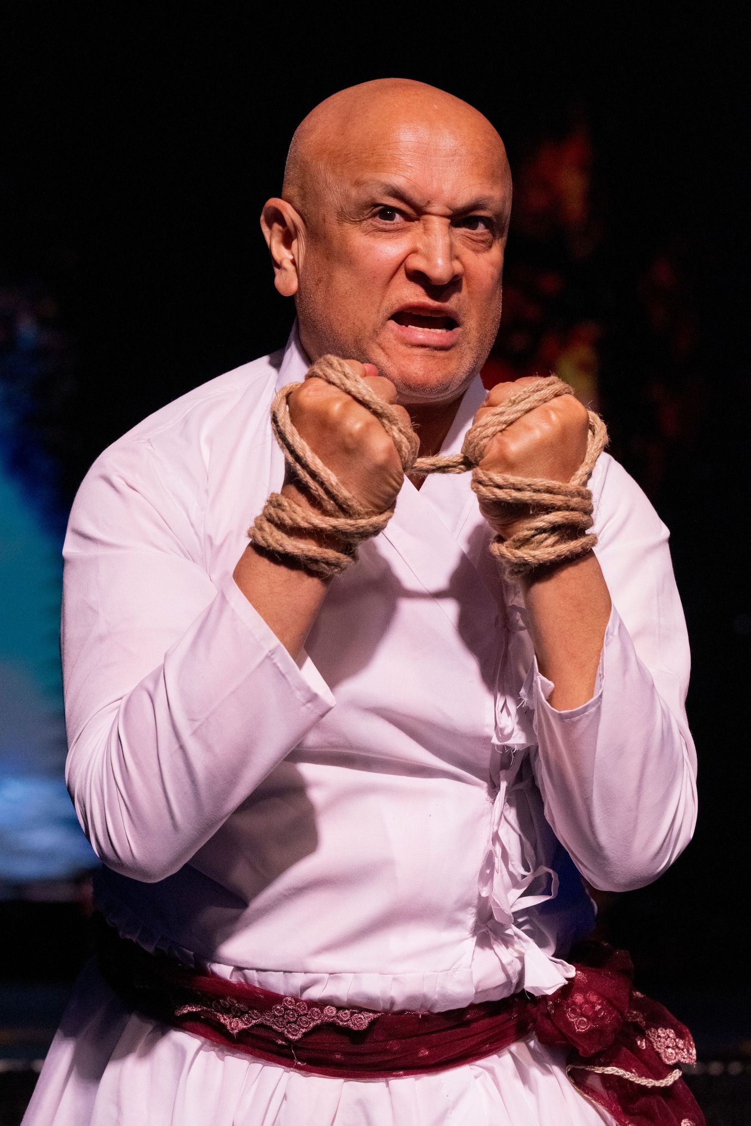Bald actor in white kurta with bound rope hands performing intense theatrical scene.