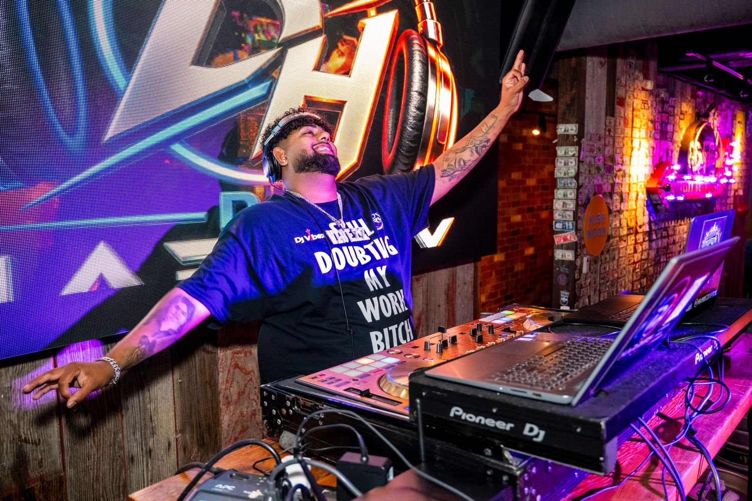 Energetic DJ performing at nightclub event wearing blue graphic t-shirt with arms raised.