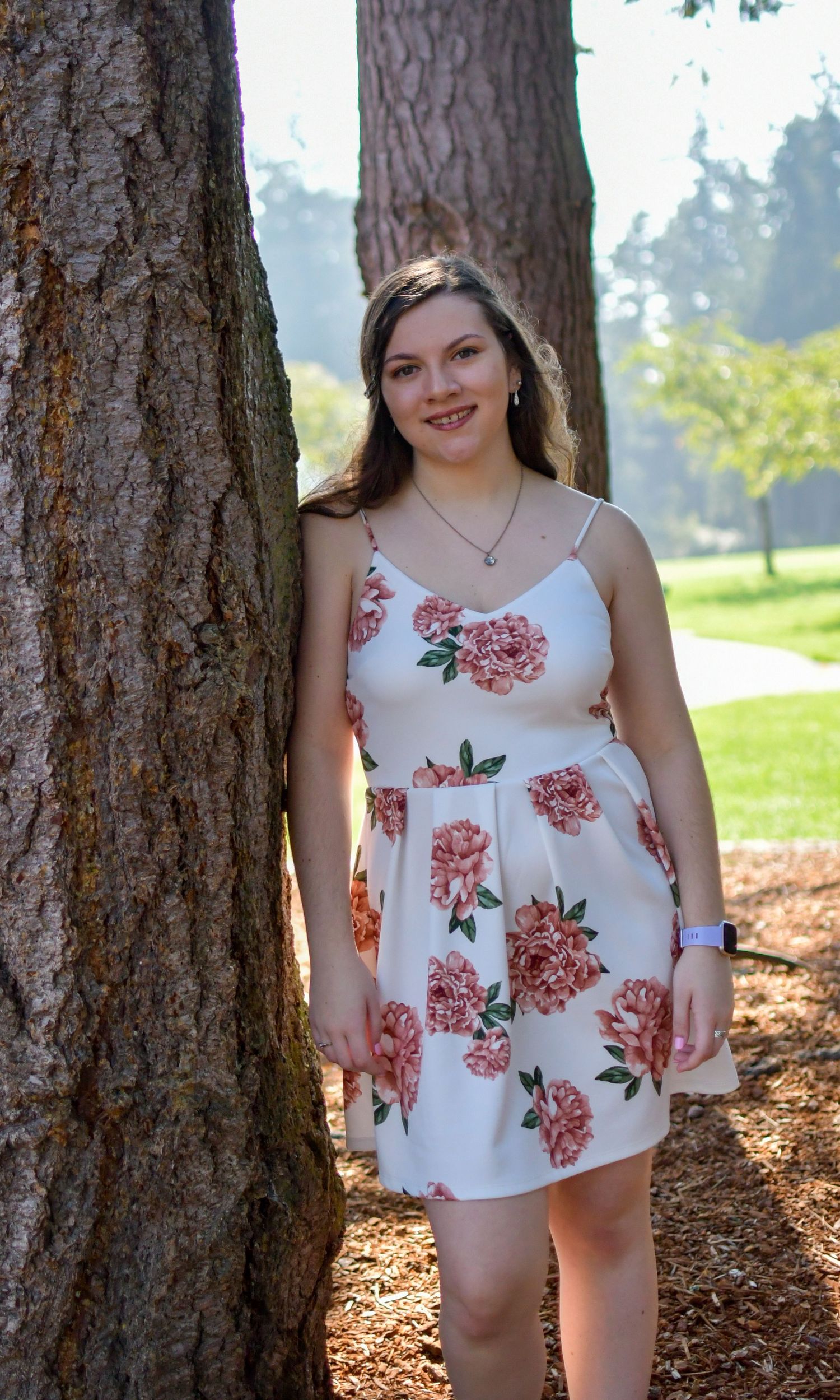 Smiling young woman in white floral sundress poses beside a large tree trunk in a sunny park.