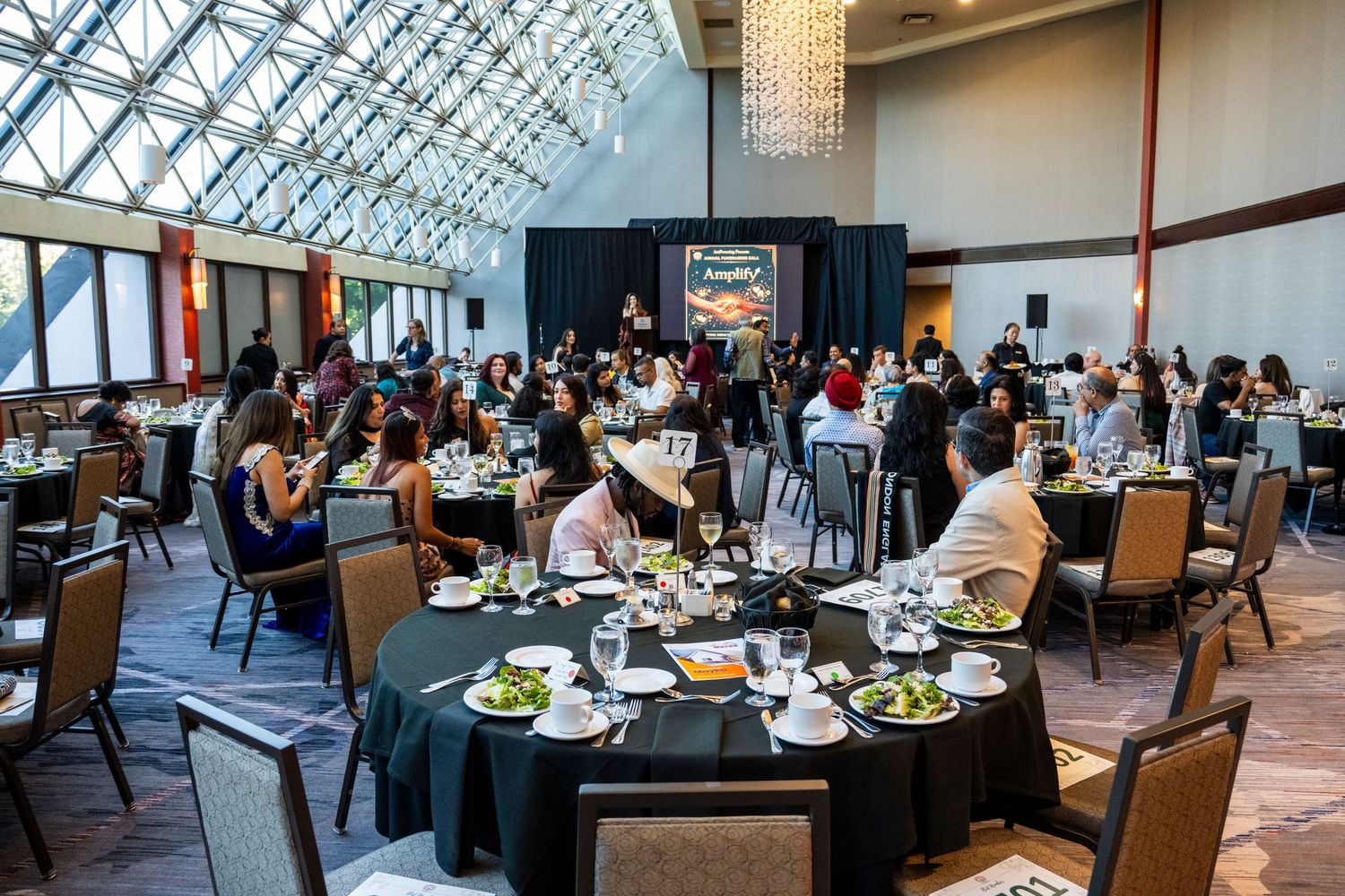 Guests enjoy a formal luncheon in a sunlit ballroom with large glass ceiling and round tables.