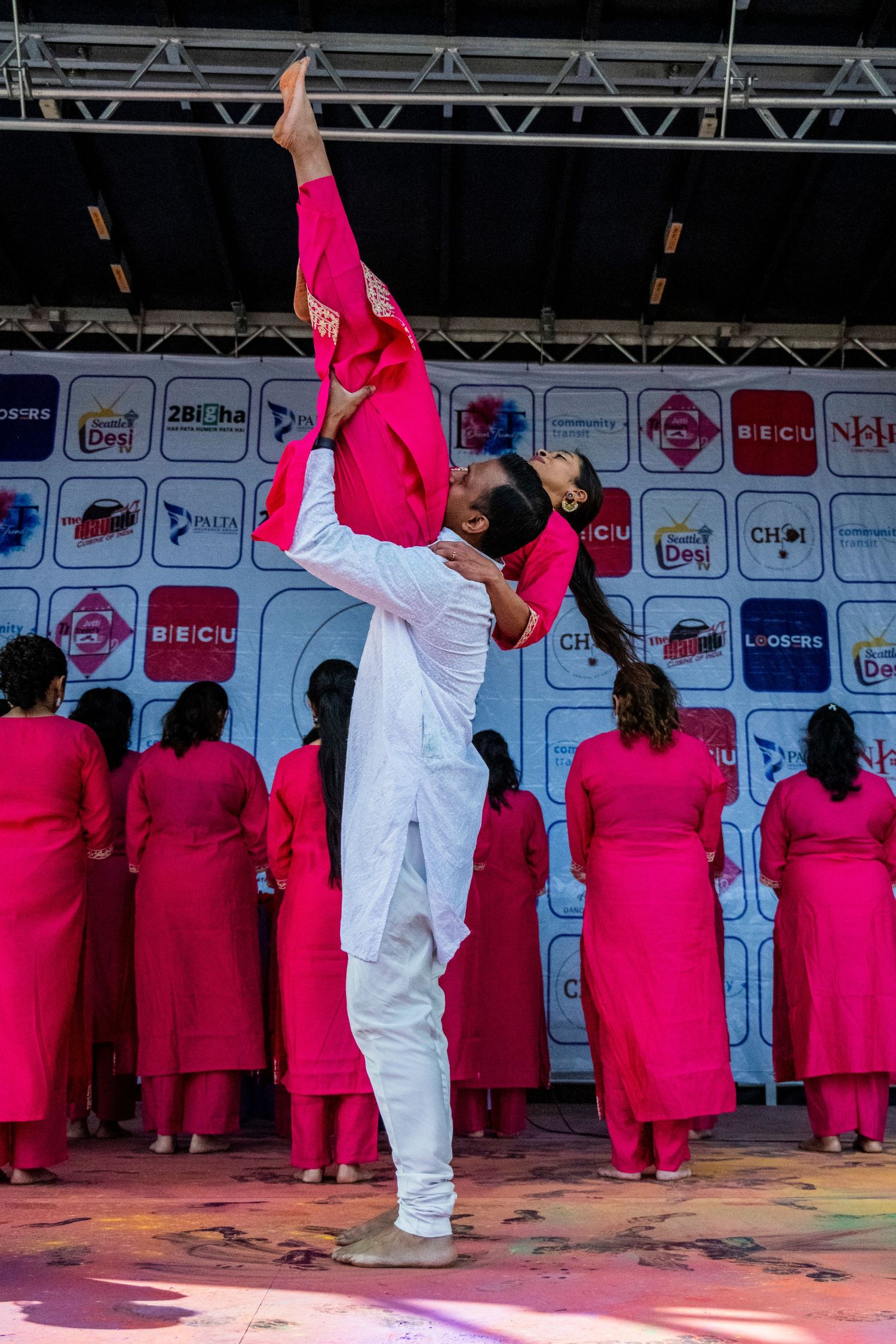 Male dancer in white lifts female dancer in pink during traditional Indian dance performance on colorful stage.
