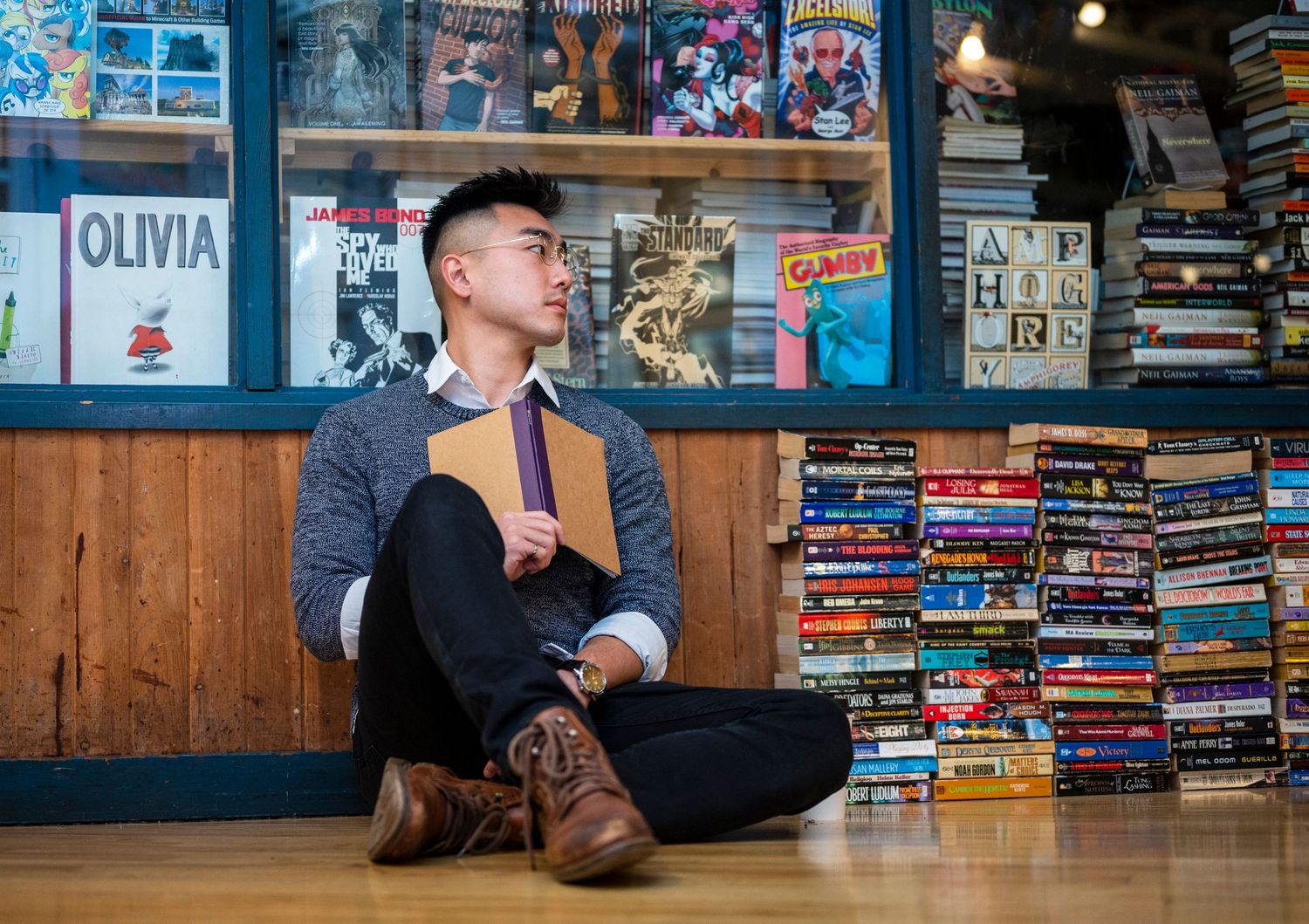 Young Asian man sitting cross-legged on a bookstore floor holding a book among stacked titles.
