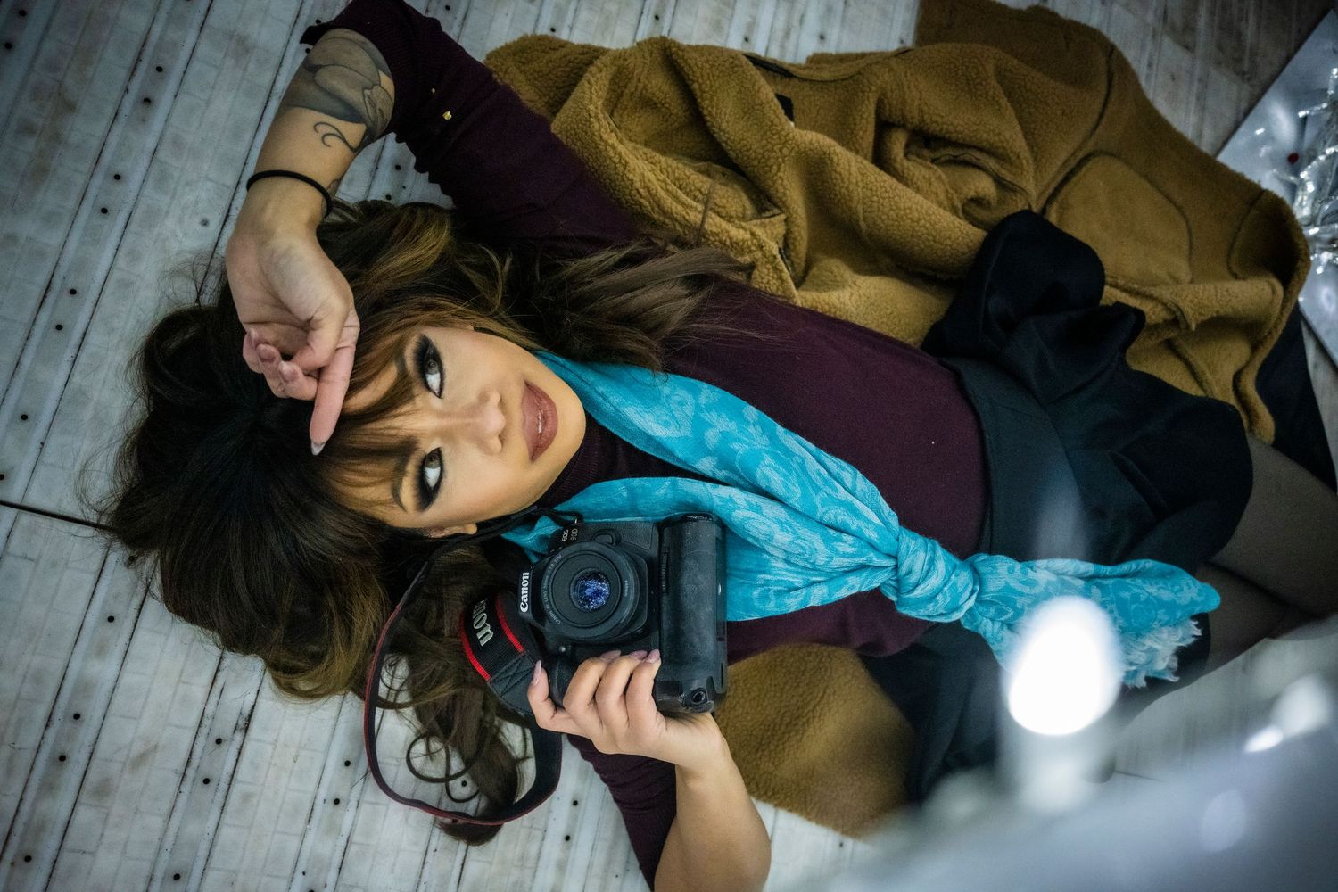 Creative woman lying on a white floor holding a DSLR camera wearing a teal scarf and coat.