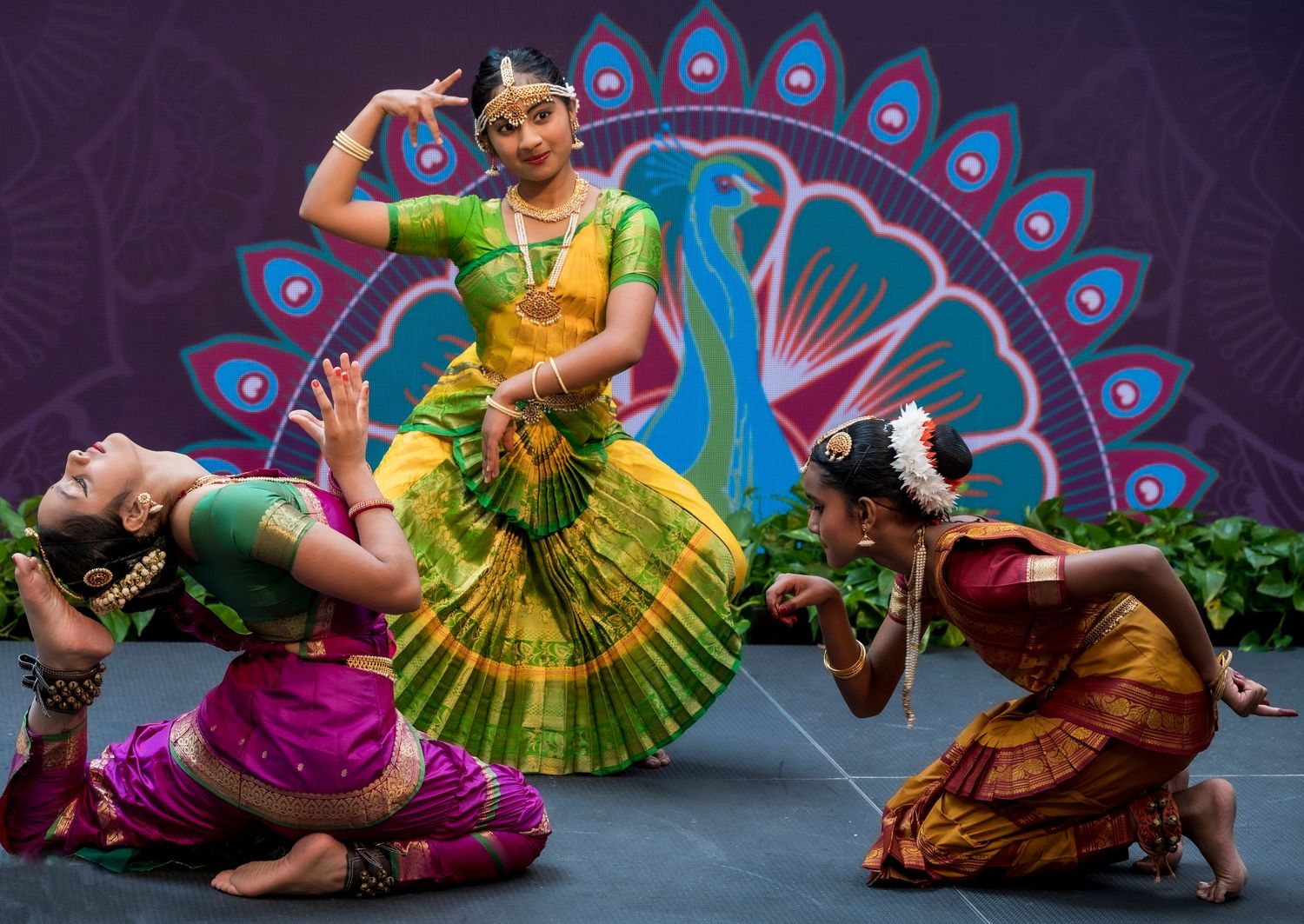 Three classical Indian Bharatanatyam dancers performing in vibrant silk costumes on a decorated stage.