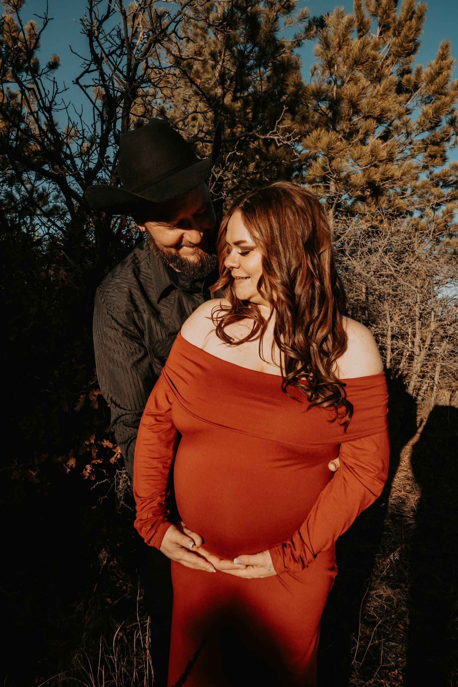 Couple maternity photo, woman in red off-shoulder dress, man in black cowboy hat at sunset.