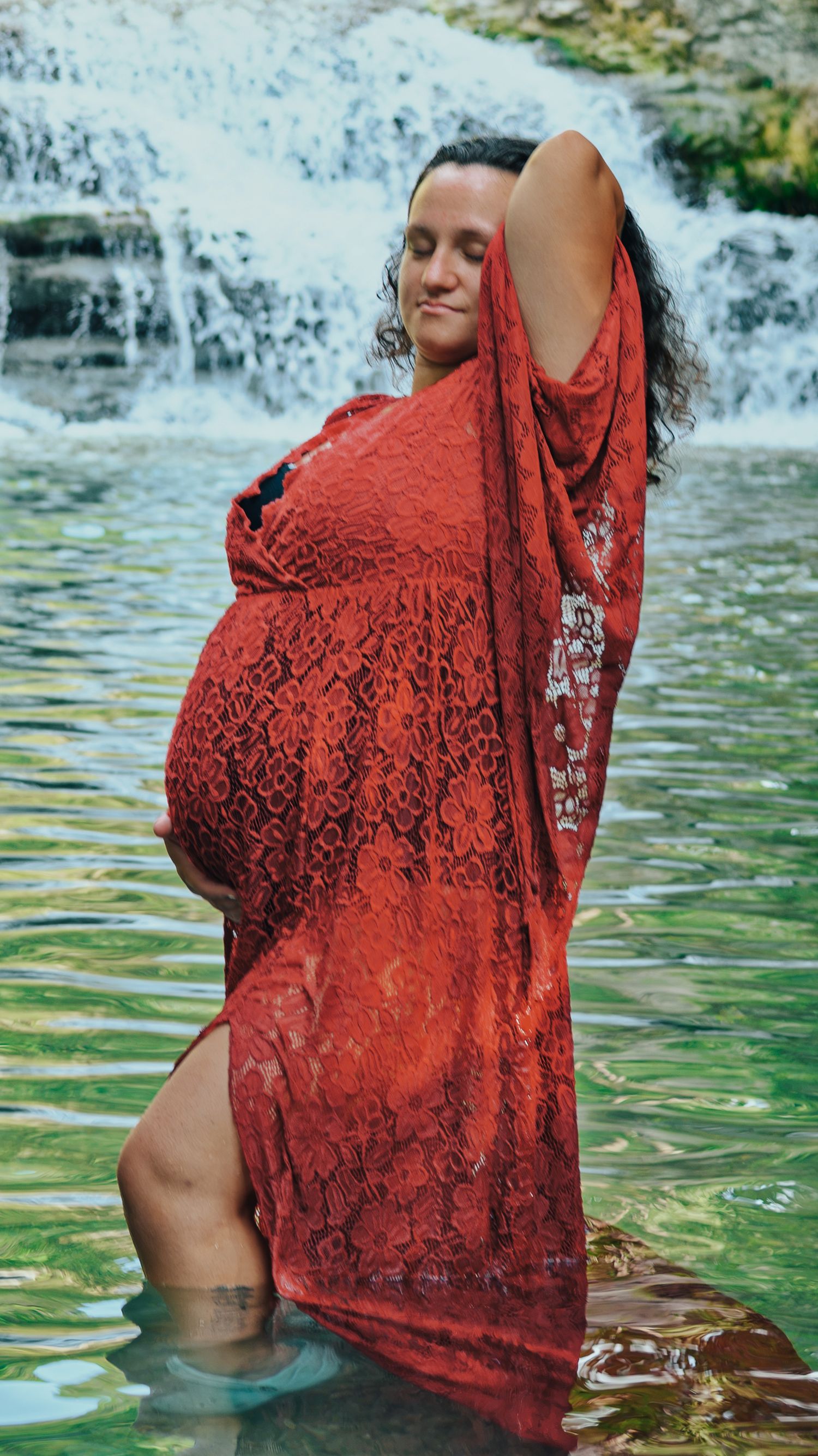 Pregnant woman in red lace dress standing in water before a scenic waterfall with eyes closed.