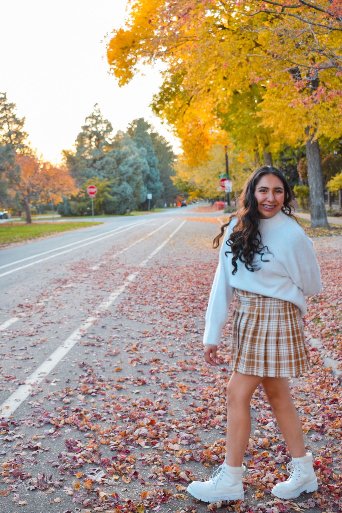 Smiling young woman in white sweater and plaid skirt standing on leaf-covered road in fall park for senior photos.