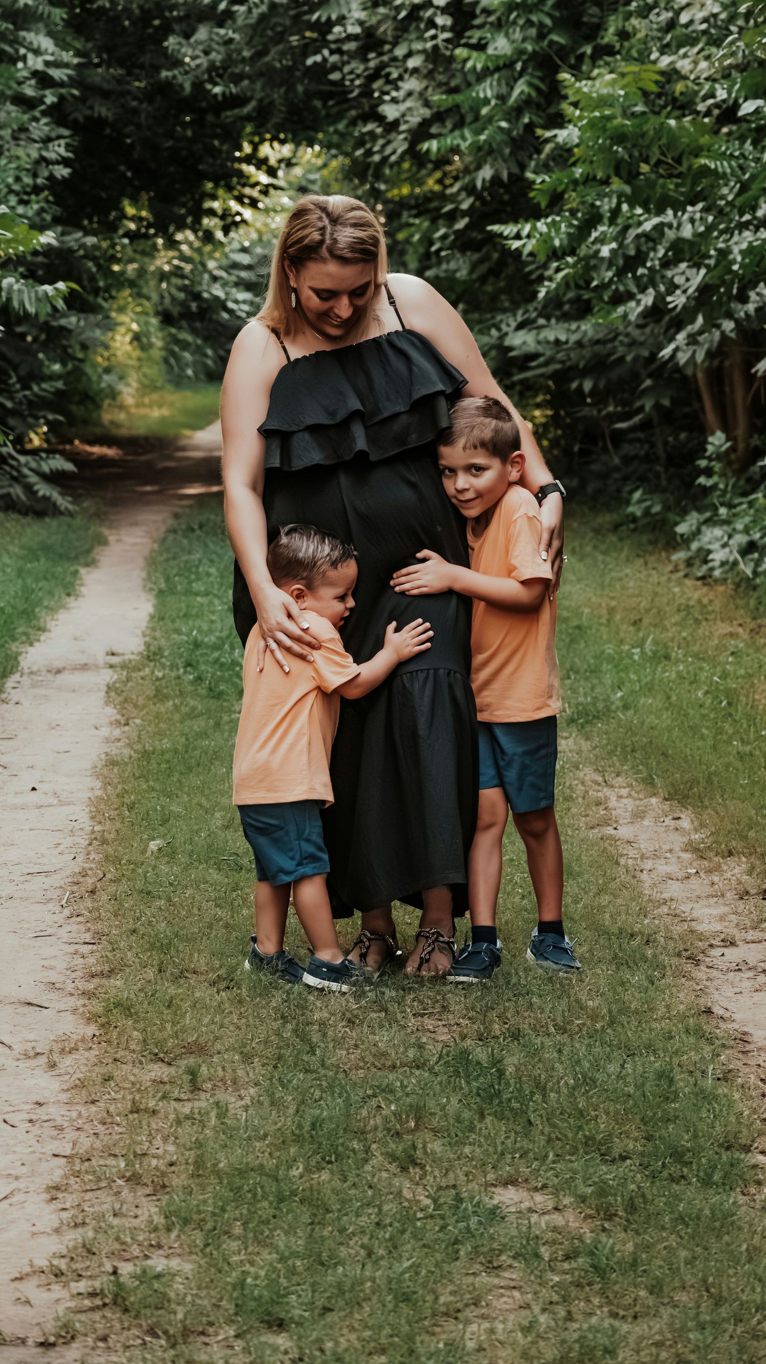 Pregnant woman in black dress hugging two young boys in orange shirts on a tree-lined path outdoors.