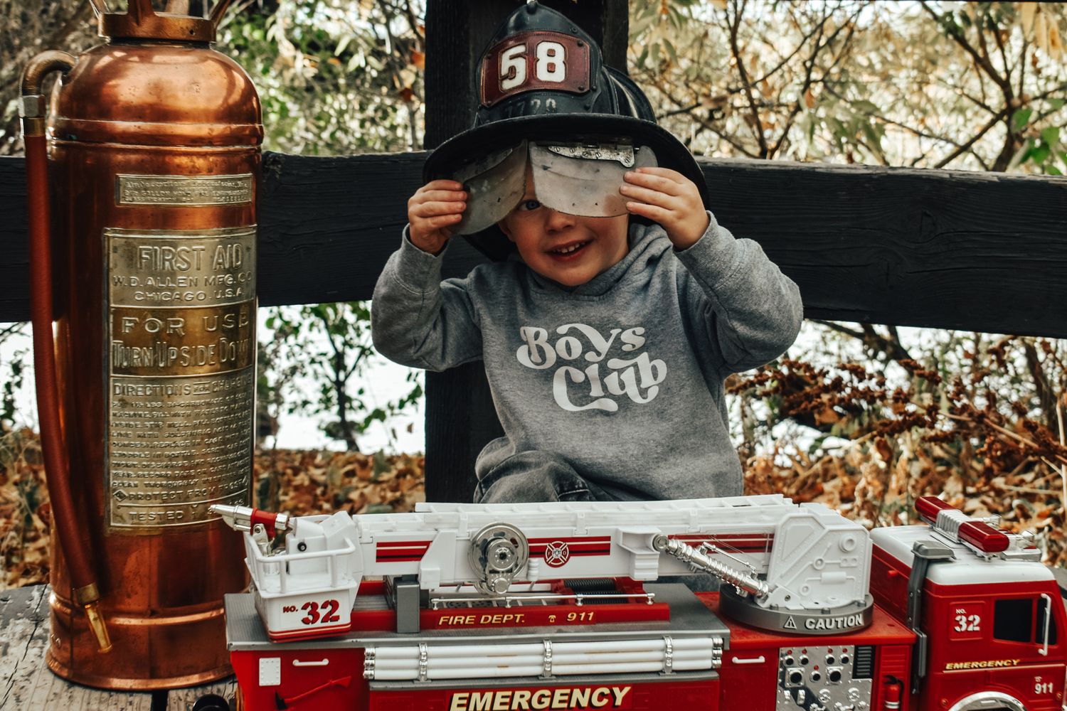 Young boy wearing a firefighter helmet playing with toy fire trucks and a vintage fire extinguisher. Family Photographer in Pflugerville, Tx
