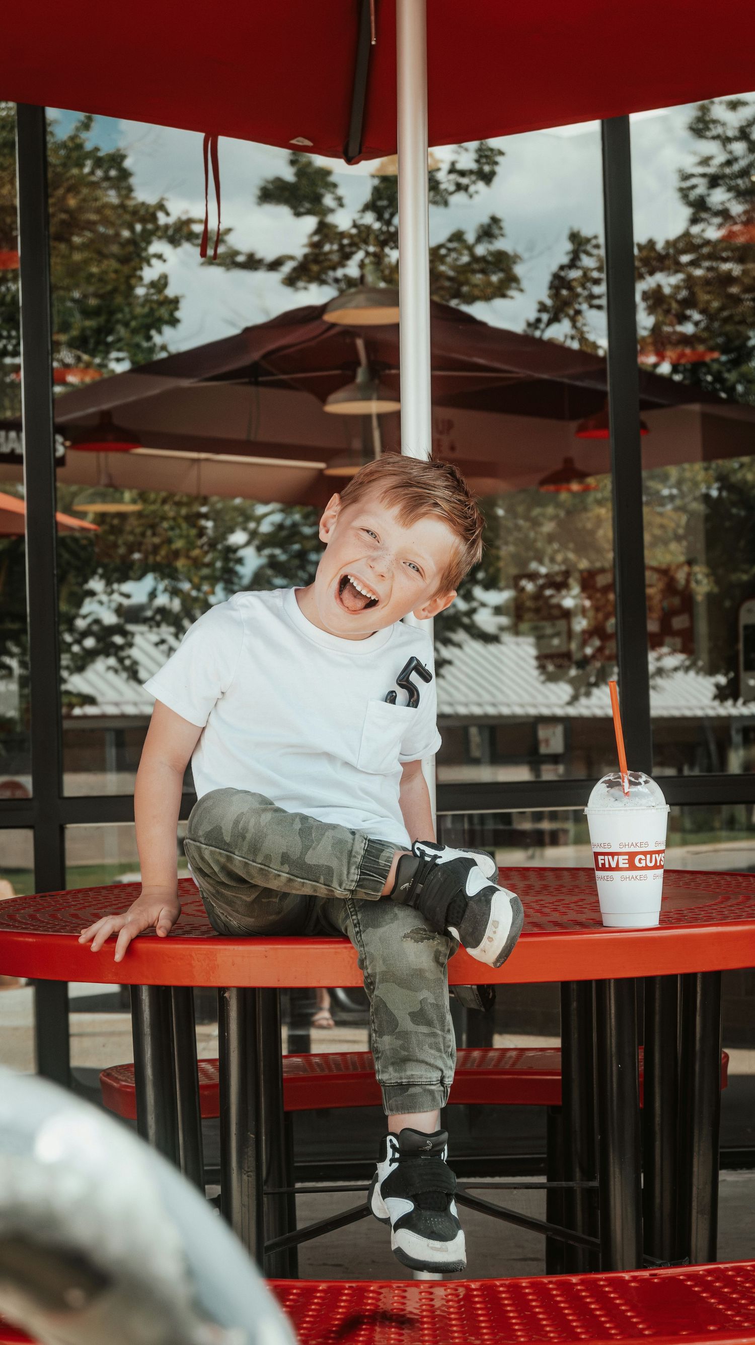 Happy toddler laughing on red outdoor table at Five Guys restaurant with milkshake nearby. Family Photographer in Pflugerville, Tx