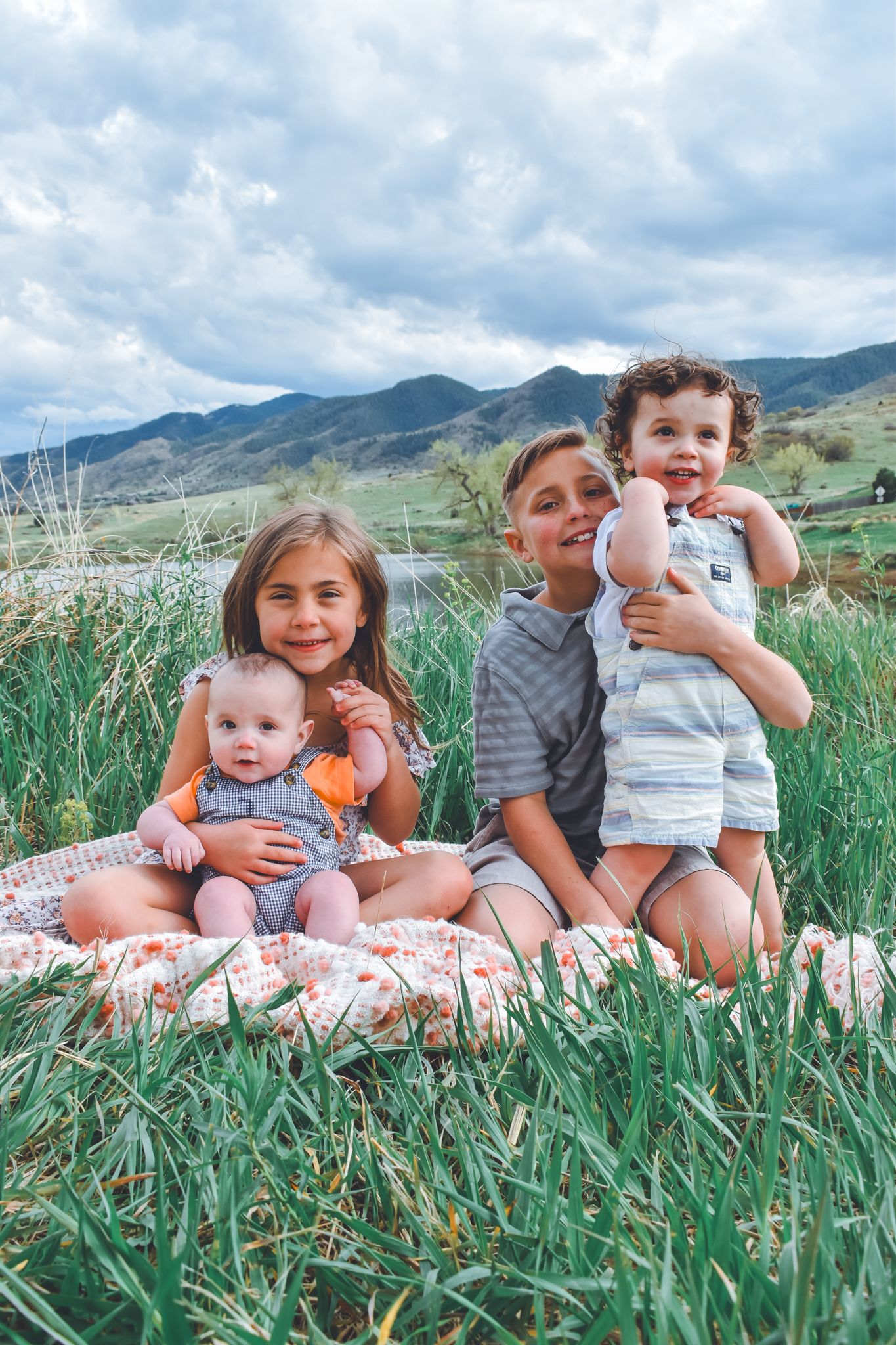 Four siblings sit together on a blanket in a green meadow with mountain views. Family Photographer in Pflugerville, Tx