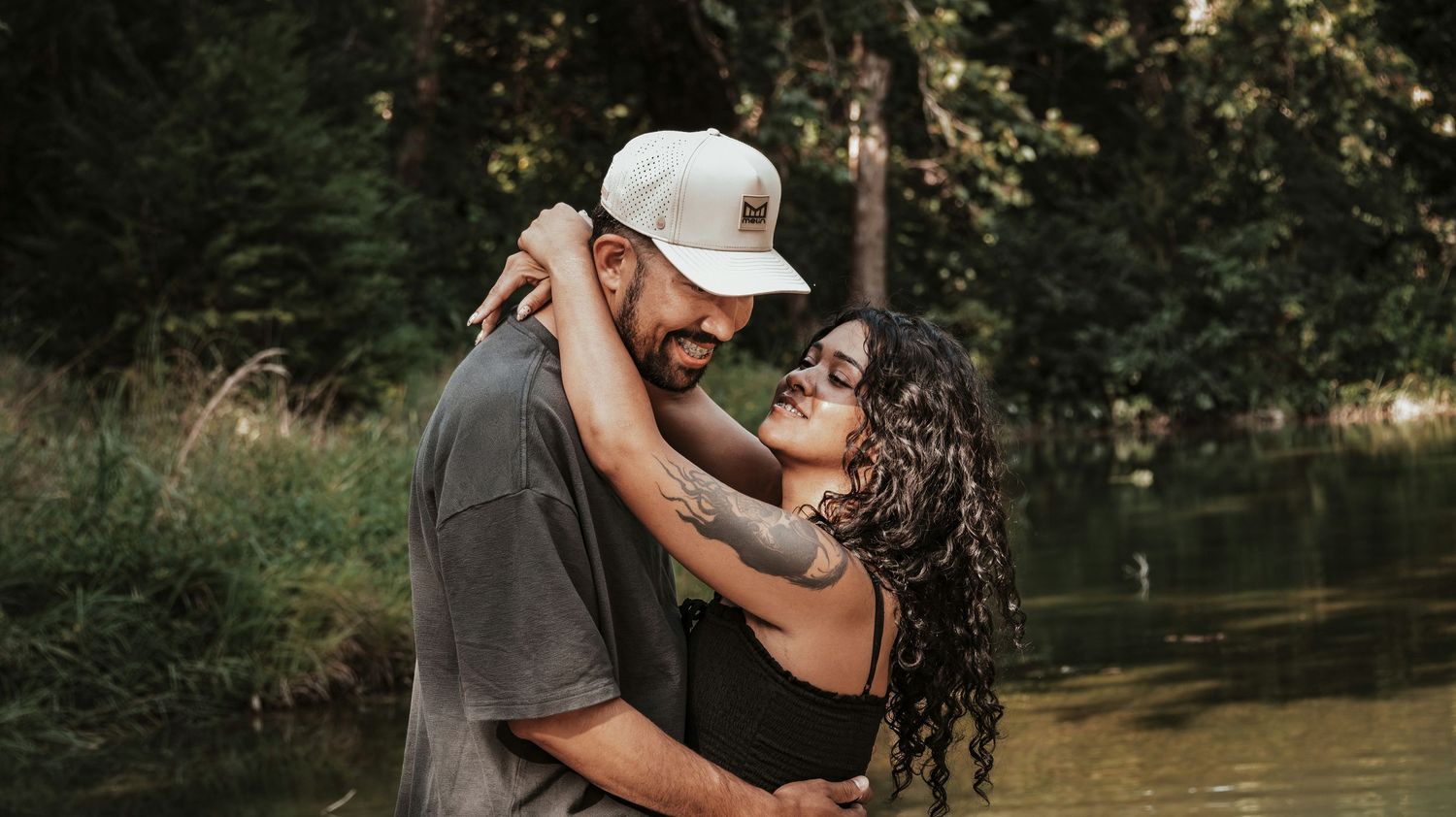 Romantic couple embracing and smiling near a peaceful forest lake during an outdoor photo session.