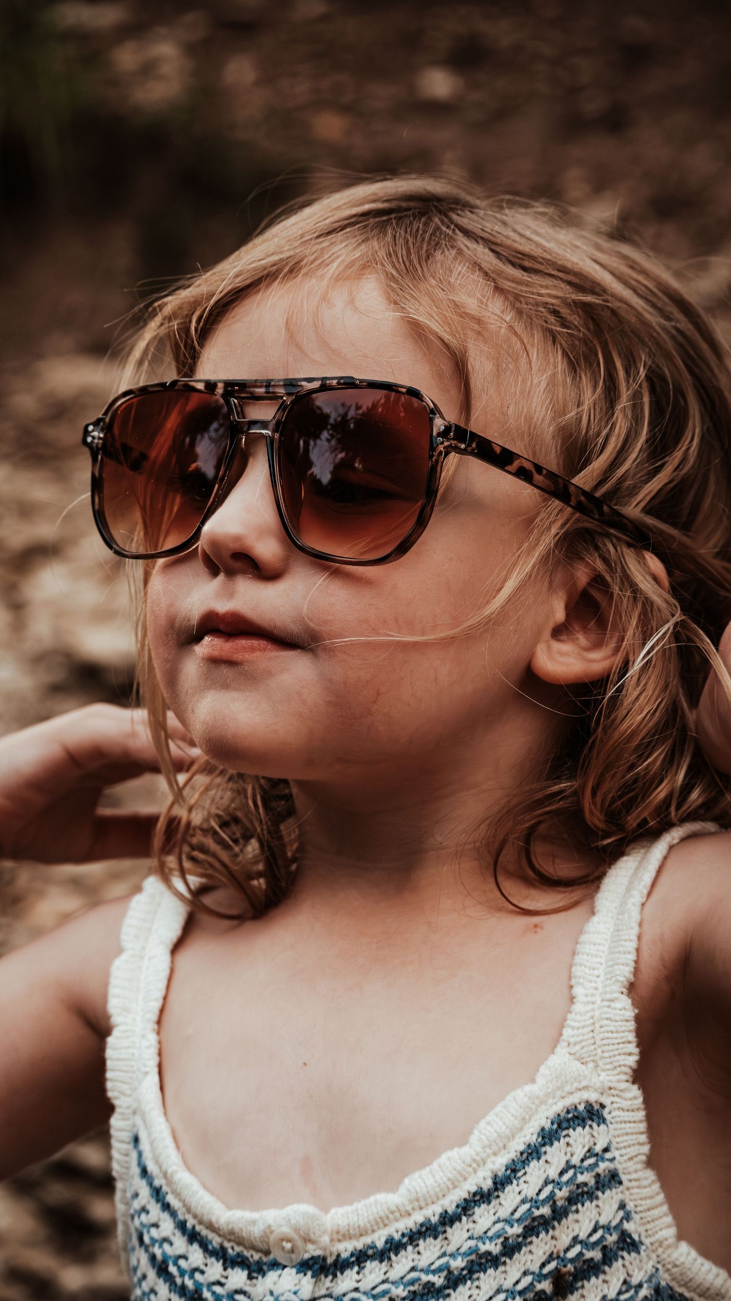 Adorable young girl posing in large tortoiseshell sunglasses and striped swimsuit in outdoor setting.