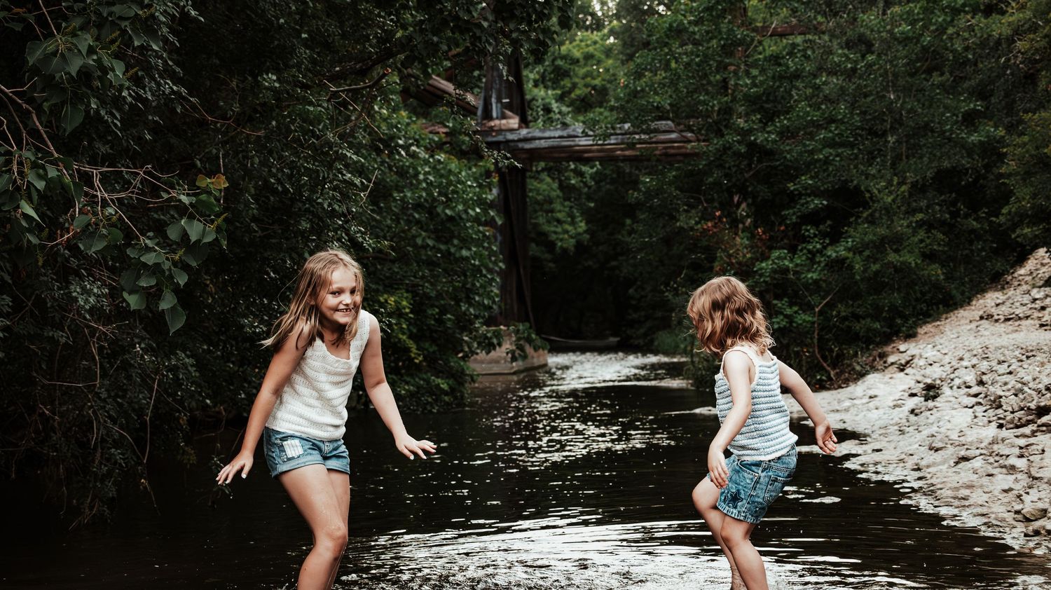 Two girls in white tank tops and denim shorts play and laugh together in a forest creek.