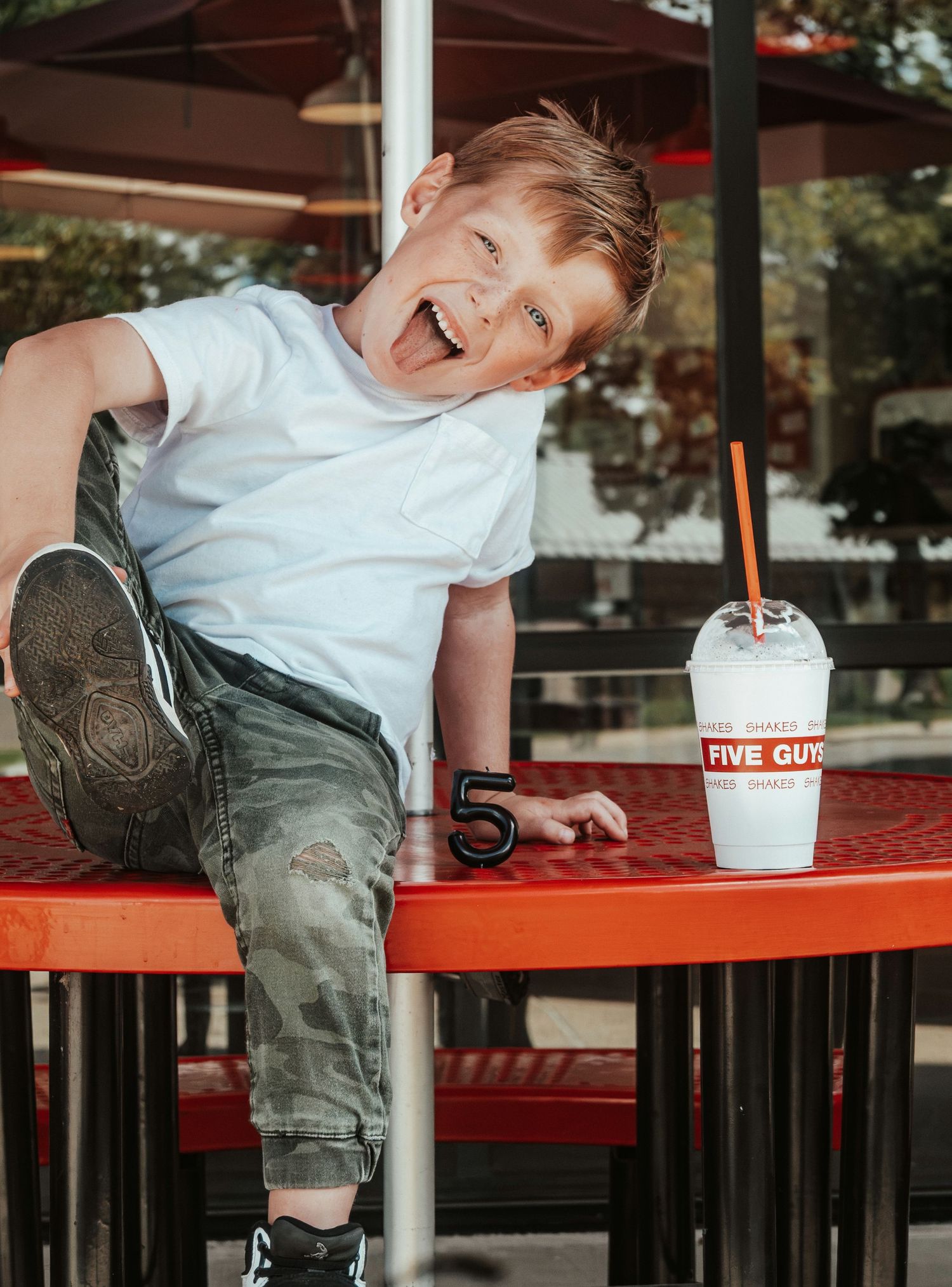 Playful boy leaning on red Five Guys outdoor table with a milkshake and number 5.