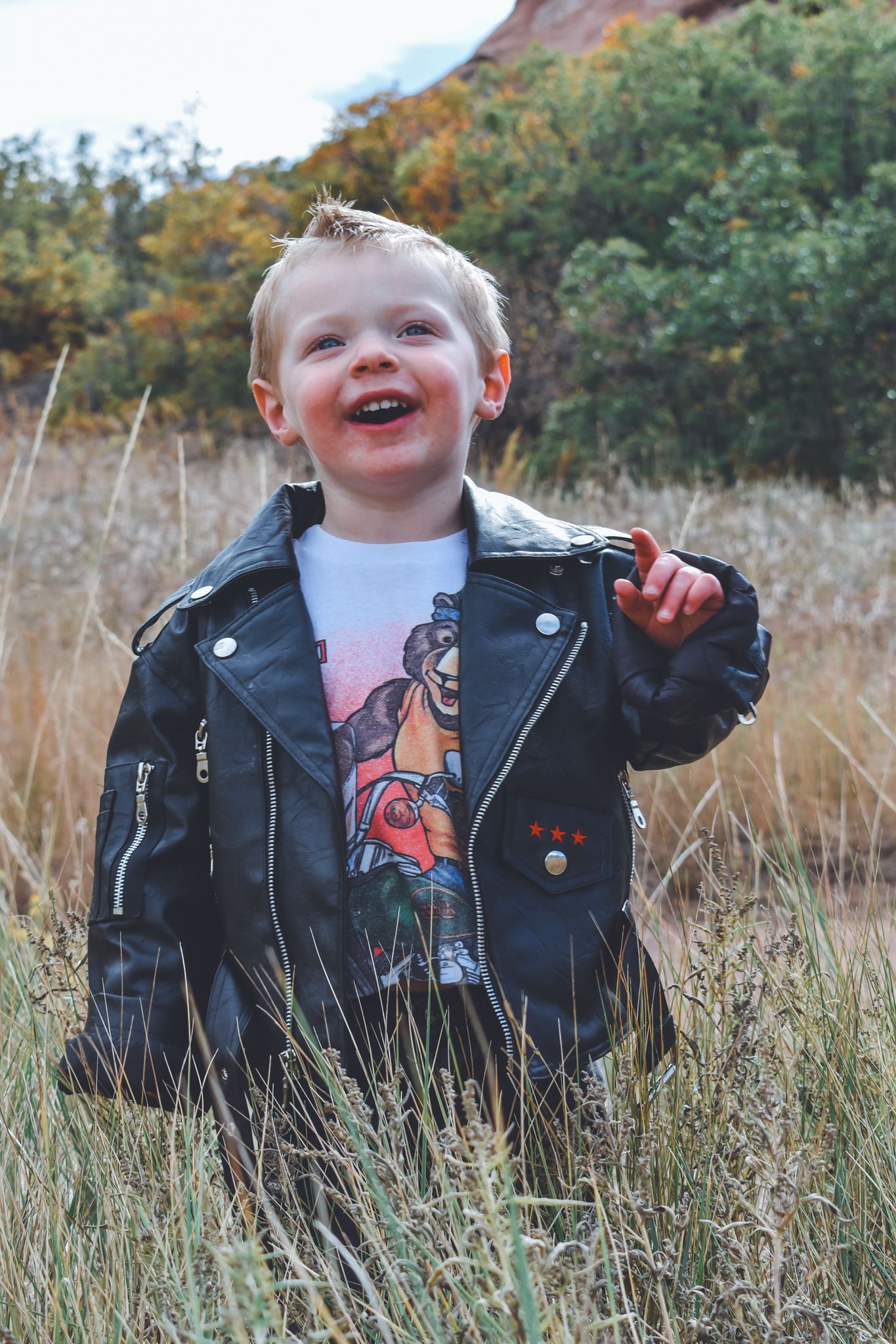 Happy laughing toddler in black leather jacket and graphic tee standing in autumn grassy field.