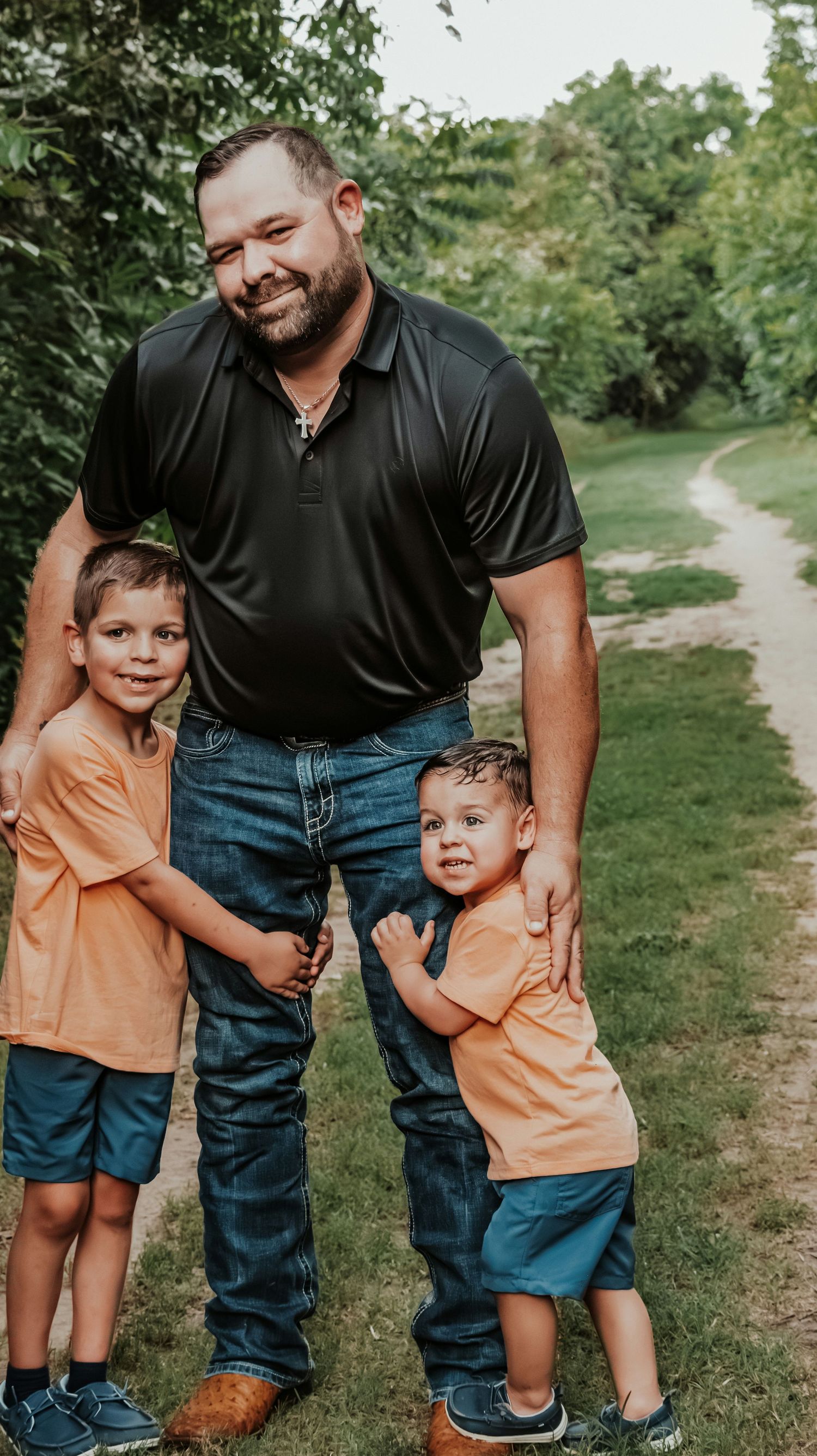 Smiling father in black polo shirt posing with his two young sons in matching orange shirts outdoors.