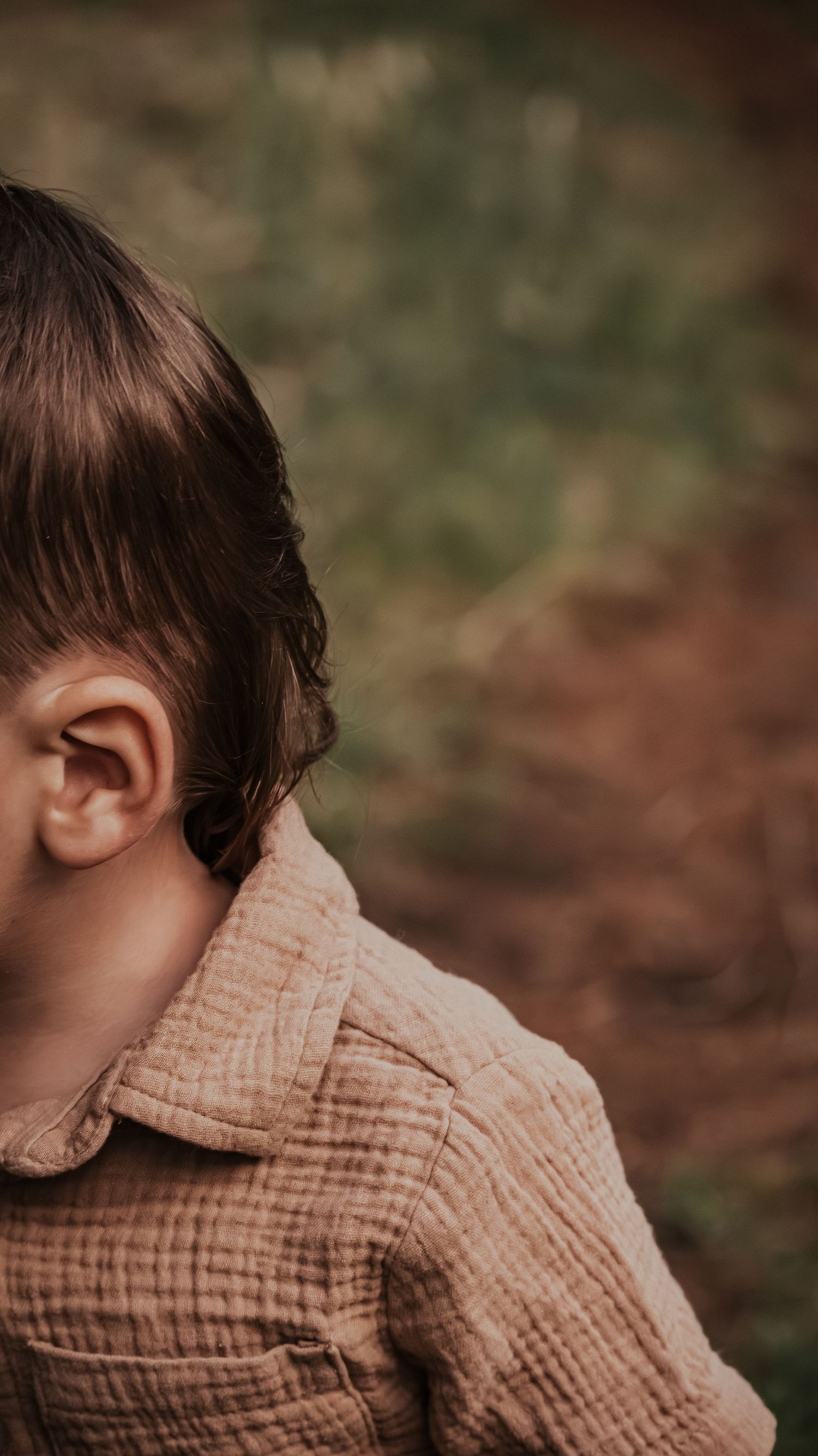 Close-up back view of a toddler boy with brown hair wearing a beige textured shirt outdoors.