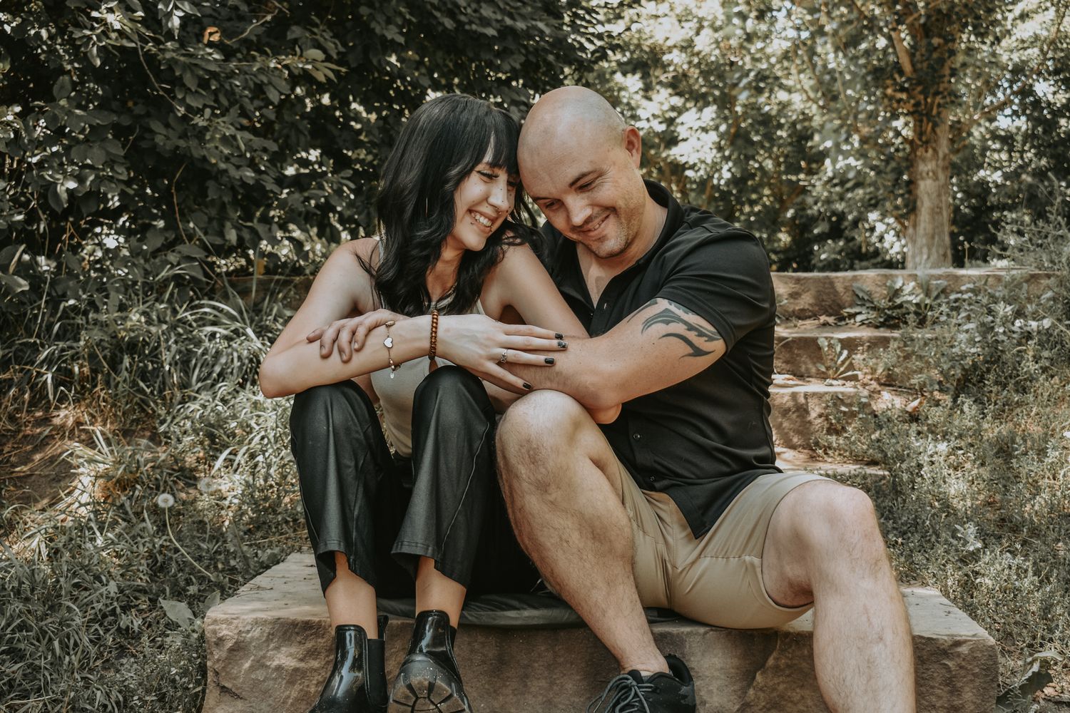 Father and daughter sits closely on a stone bench outdoors, smiling and holding hands in a lush garden setting for senior photos.