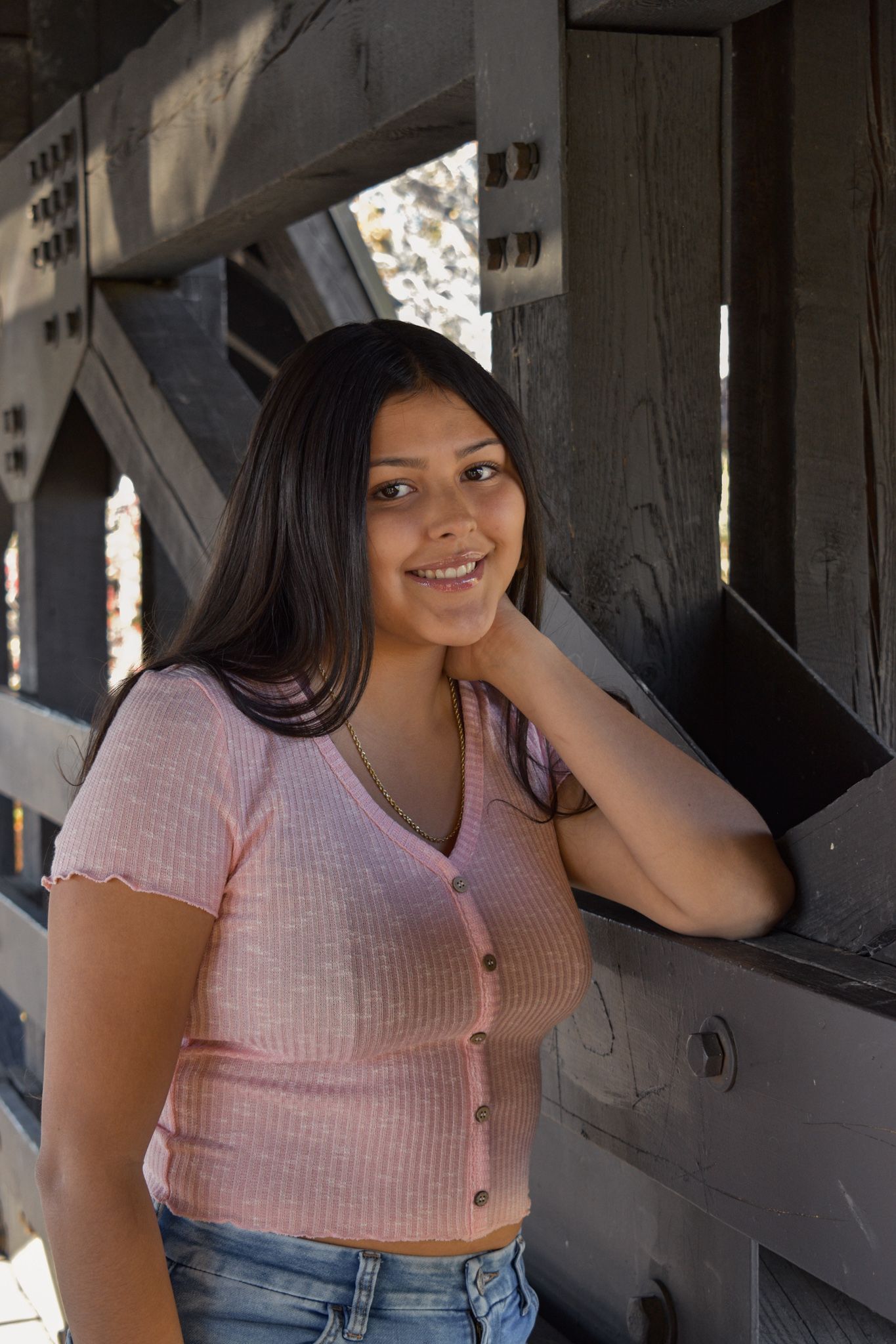 Smiling young woman in pink top leans against wooden bridge railing outdoors for senior photos.