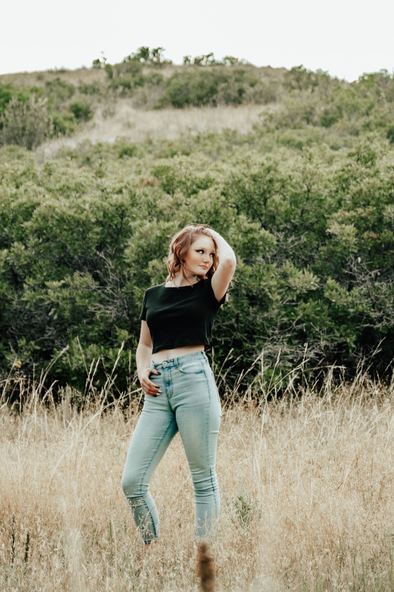 Redhead woman in black crop top and light jeans posing in dry golden grass field with green hills behind for senior photos.