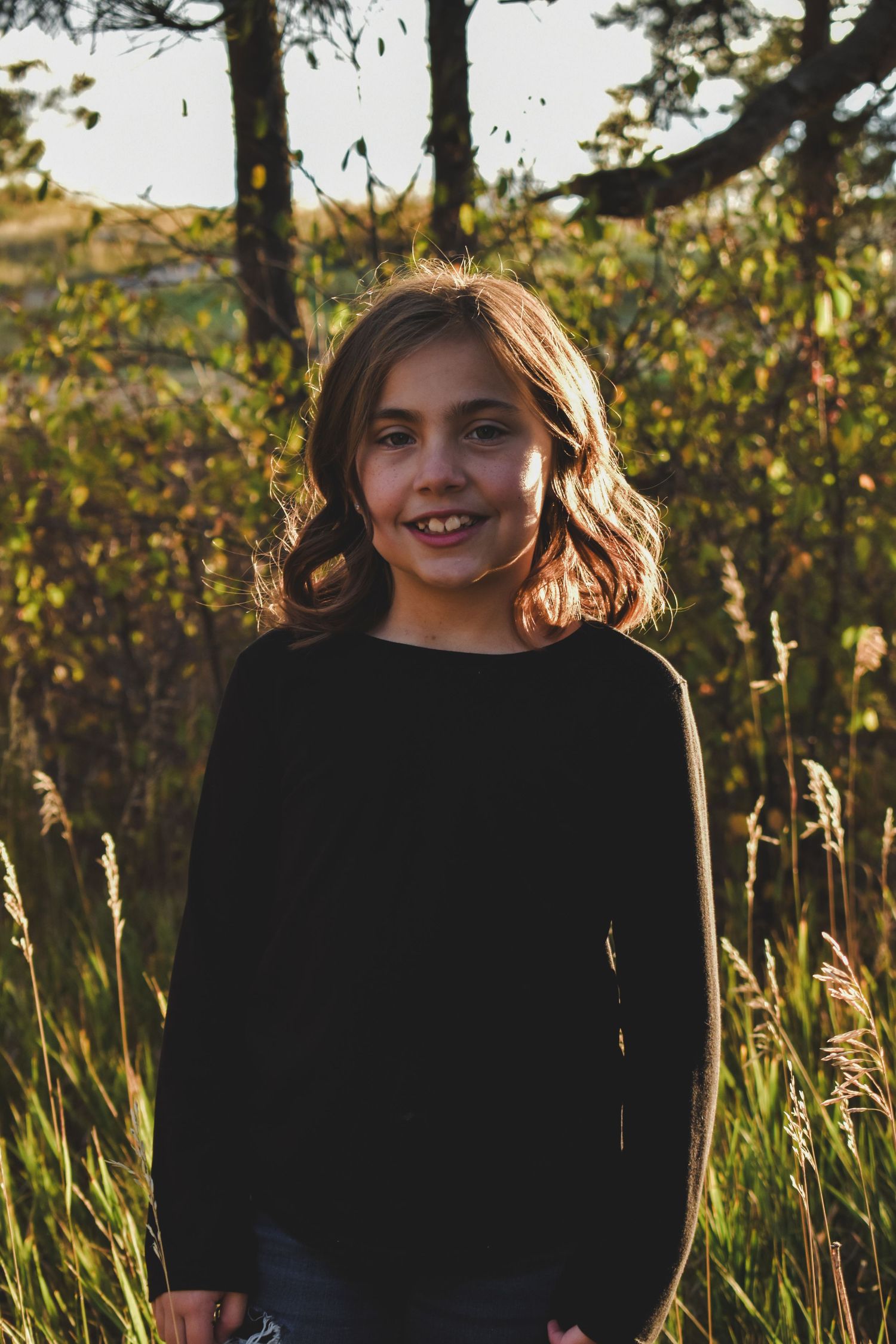 Smiling young girl in black long-sleeve shirt standing in golden sunlit field near trees.