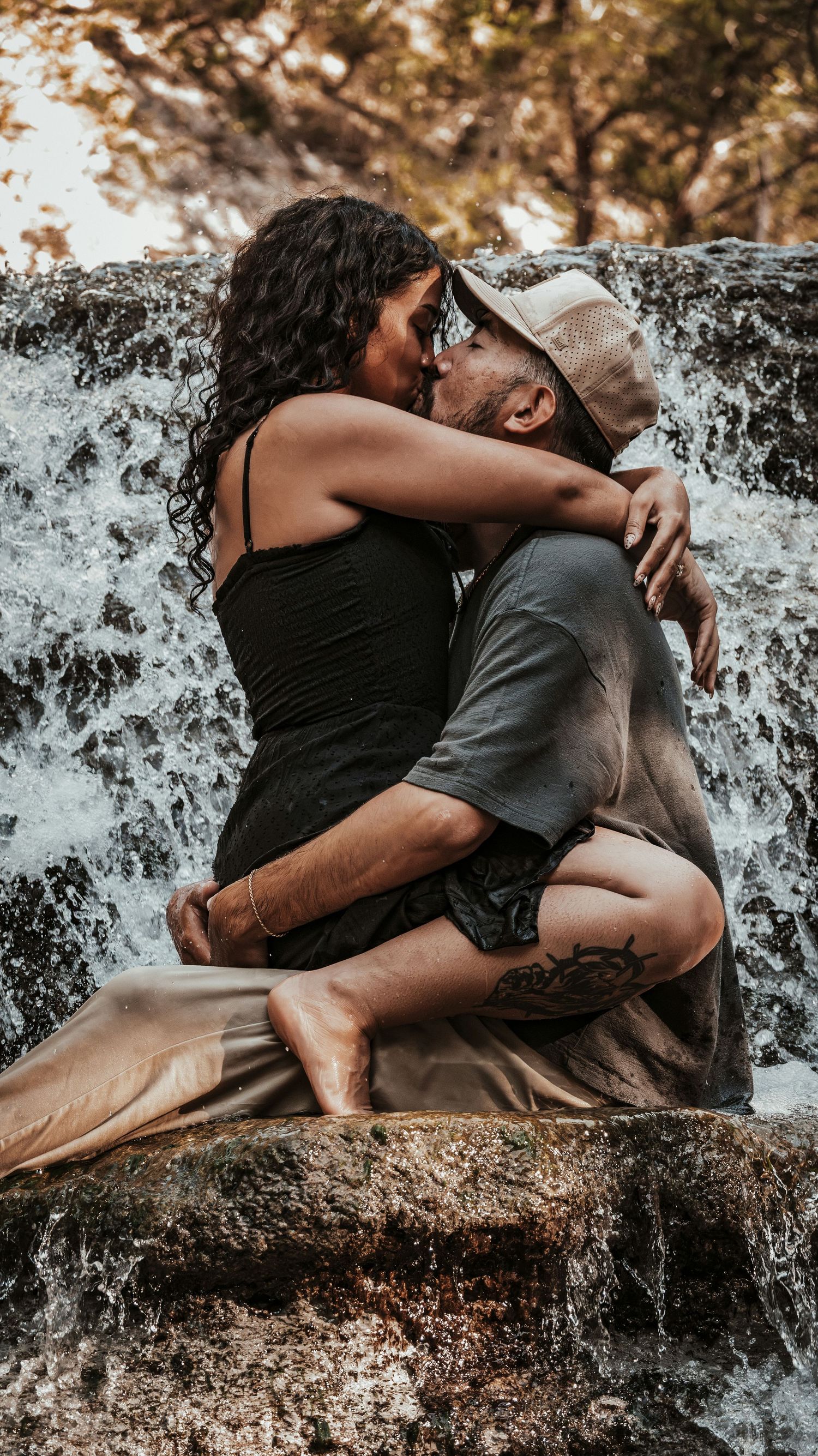 Romantic couple kissing and embracing on rocks beside a flowing waterfall outdoors.