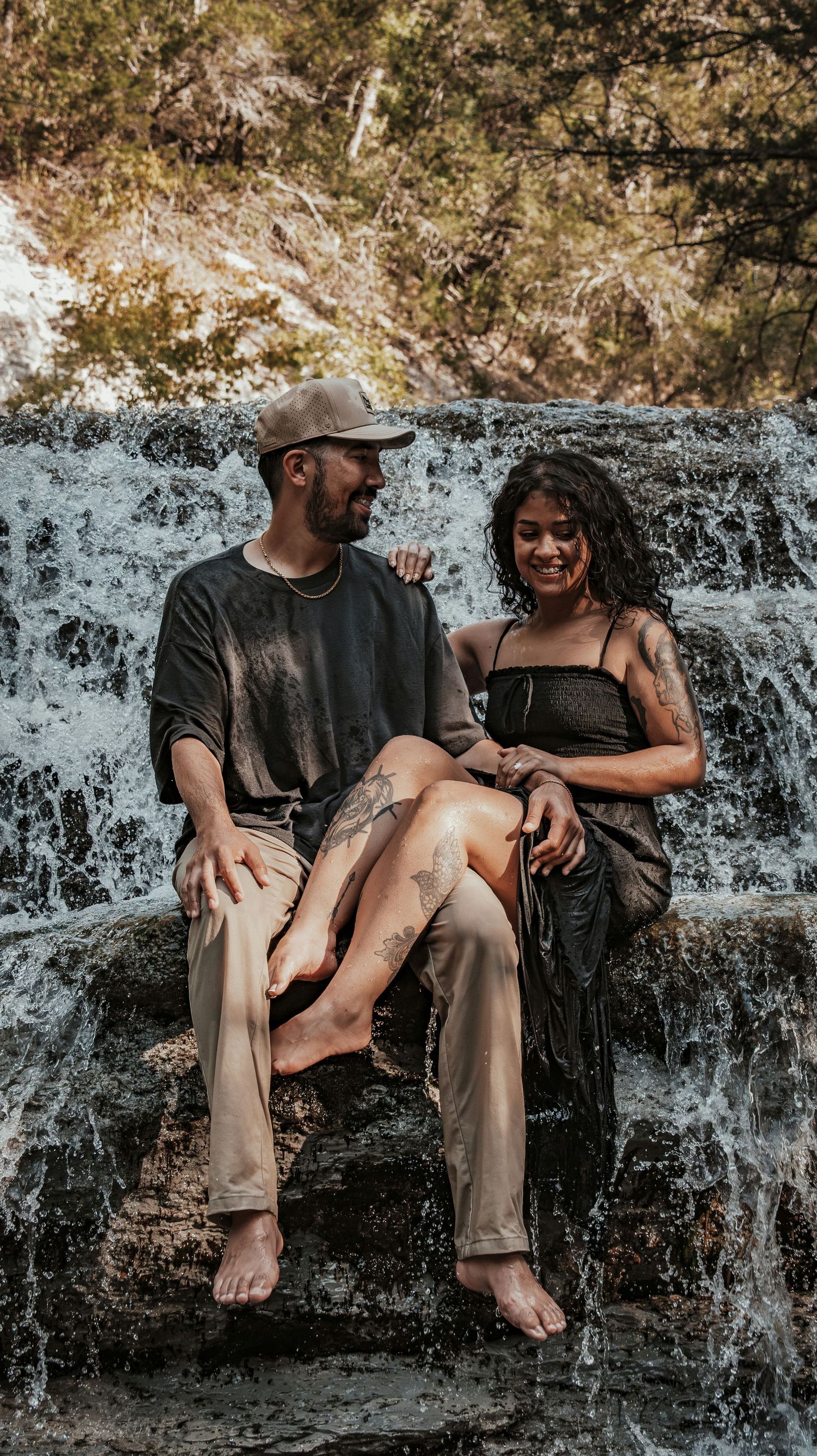 Smiling couple sitting barefoot together on rocky steps with a scenic waterfall backdrop in nature.
