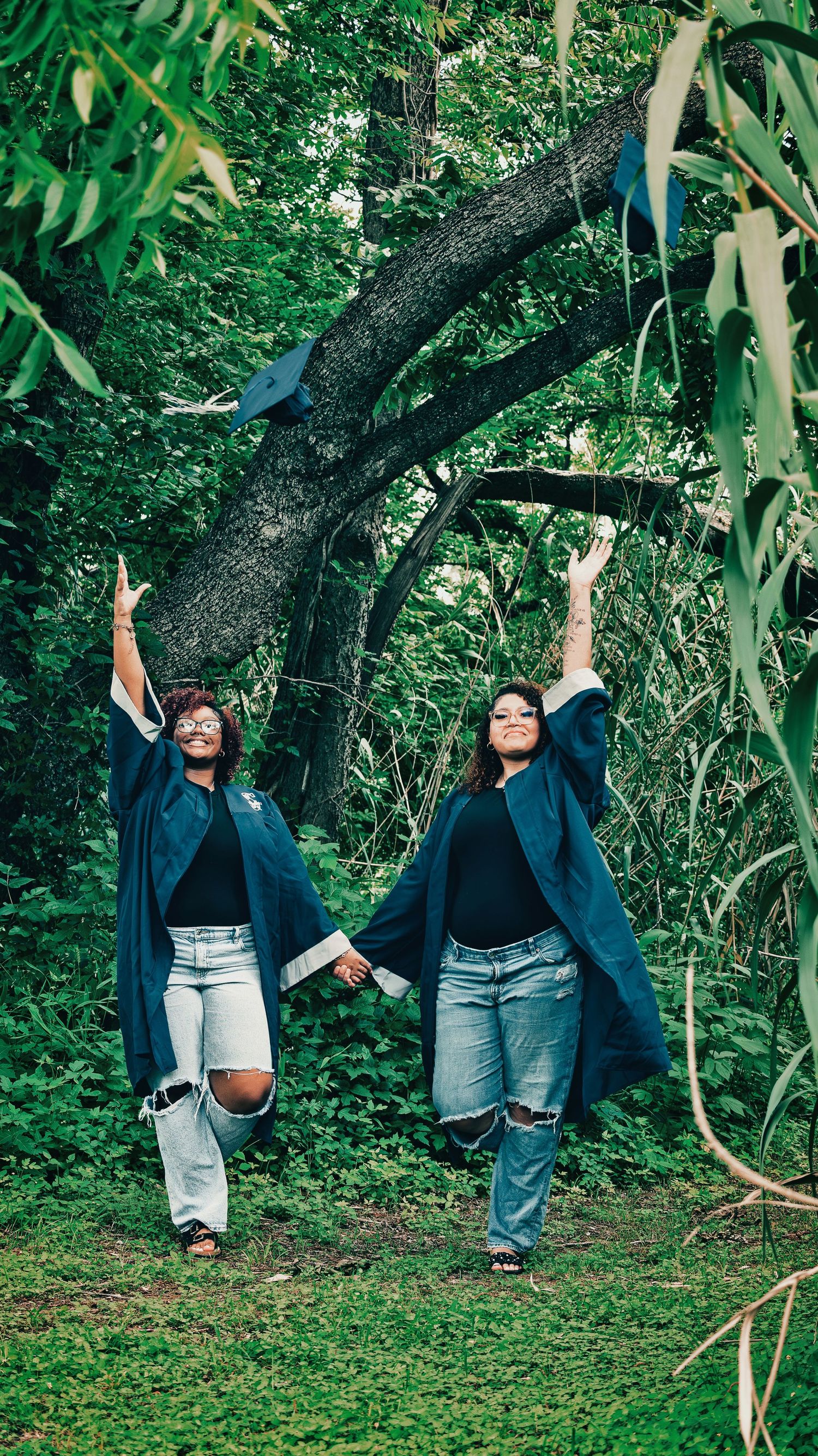 Two smiling graduates in blue gowns holding hands and raising arms in a lush green forest for senior photos.