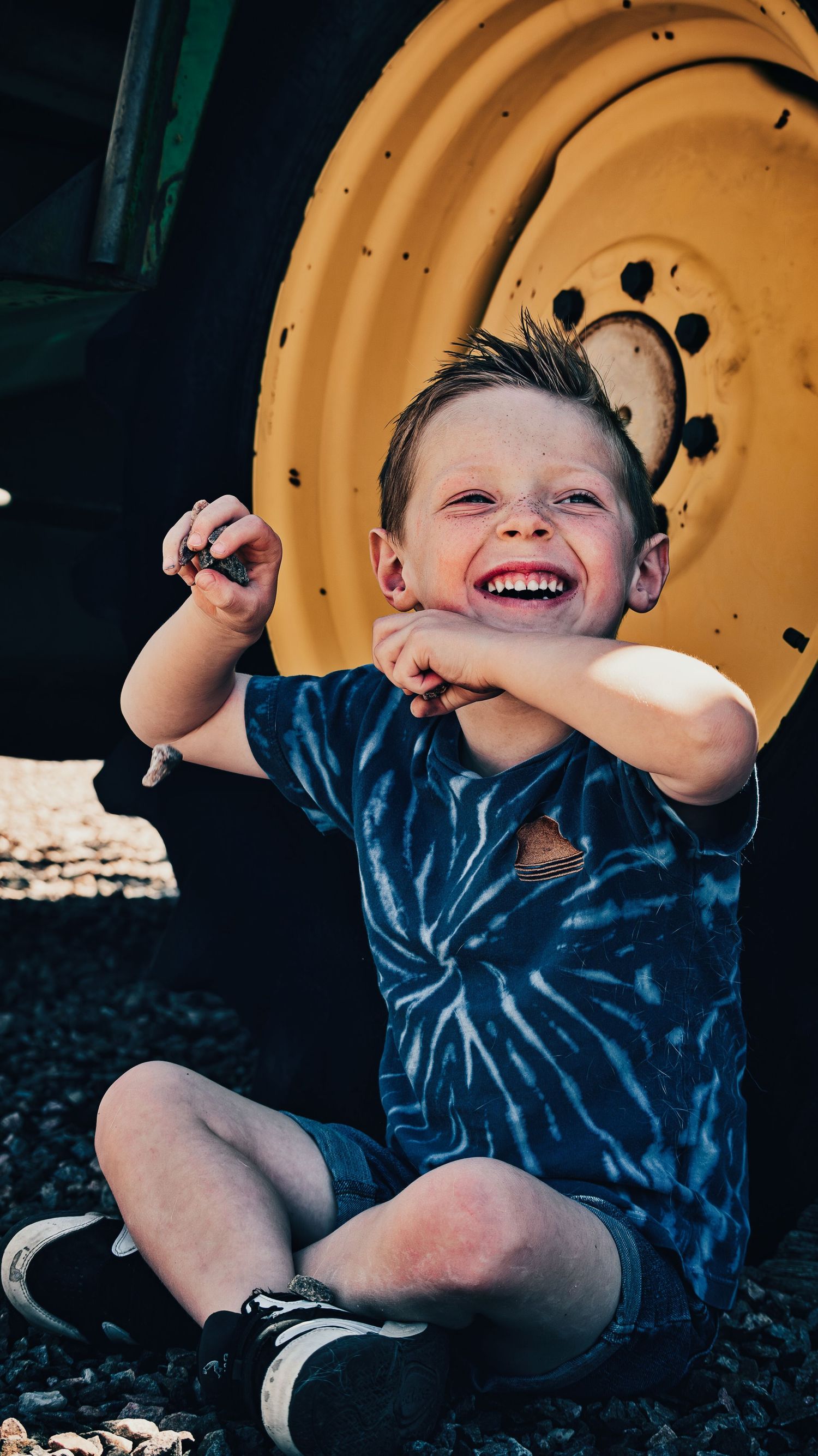 Laughing boy in tie-dye shirt sits on gravel in front of large yellow tractor tire. Family Photographer in Pflugerville, Tx